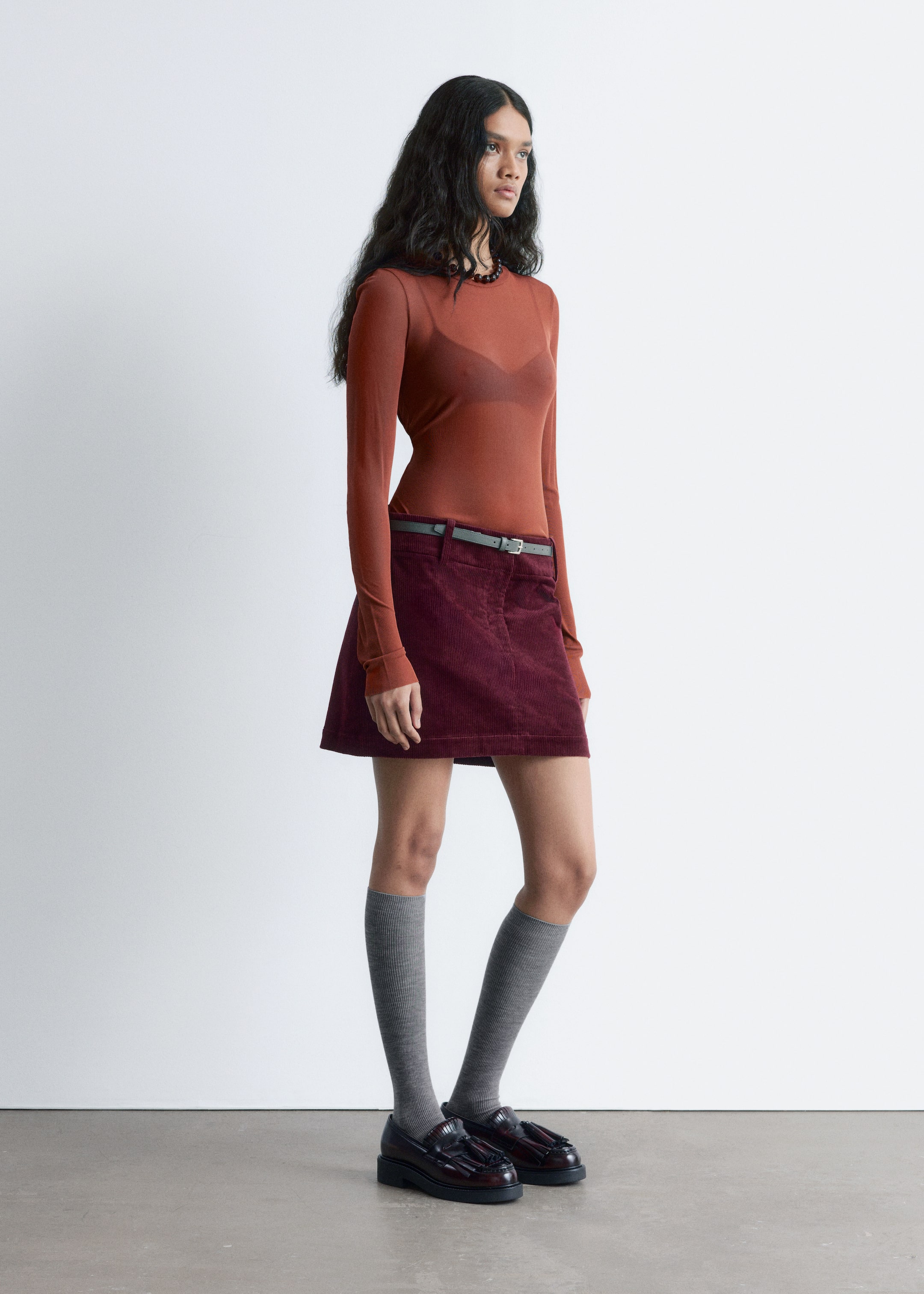 View larger image: An outfit is being presented in a side profile, featuring a sheer rust-colored long-sleeved top with a round neck, layered over a dark bra, and paired with a burgundy corduroy A-line mini skirt. The skirt is cinched by a thin grey belt with a silver buckle, and the ensemble is completed with grey knee-high socks and dark red patent leather loafers with tassels and a chunky sole.