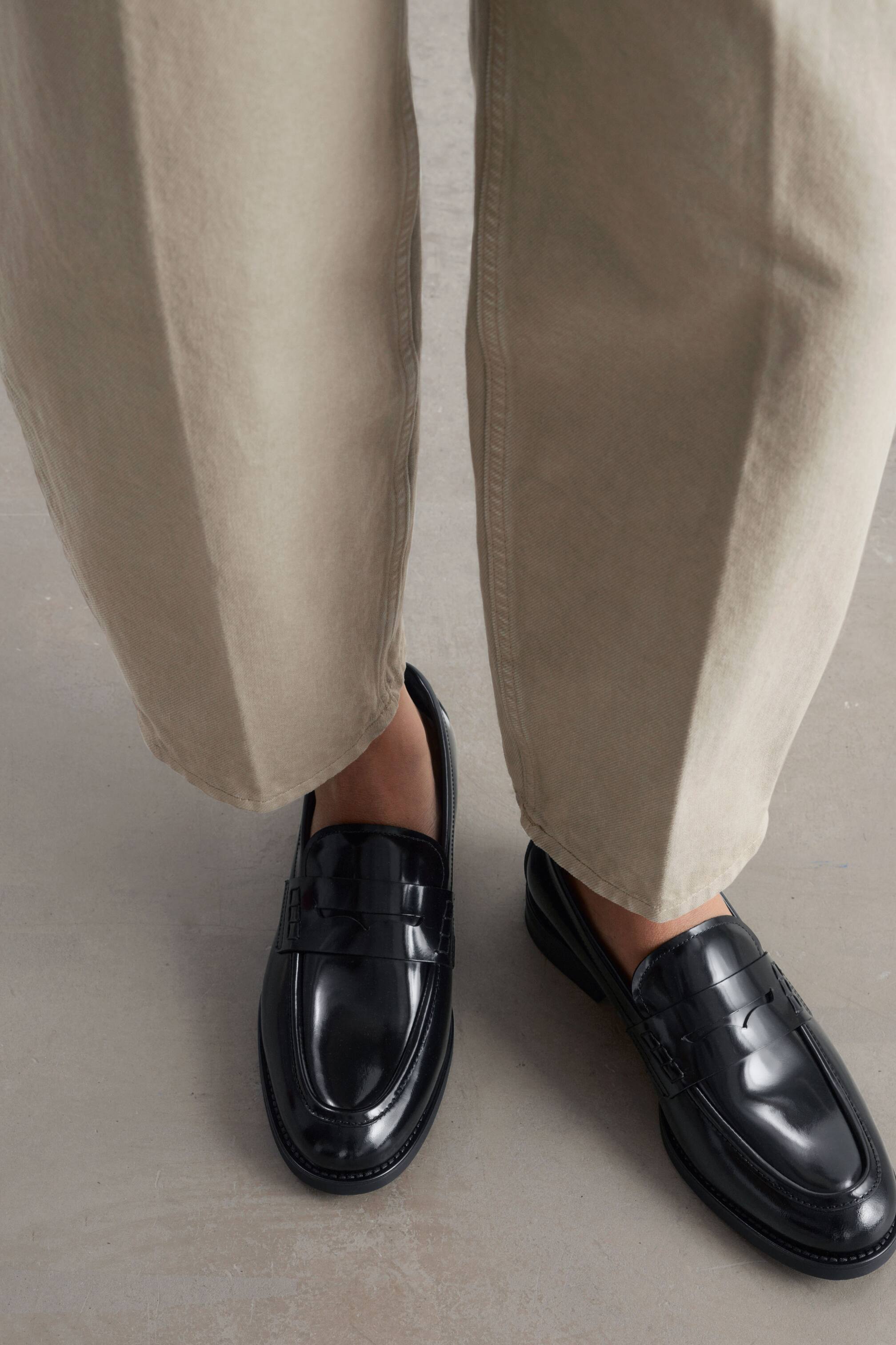 Zobrazit větší obrázek: Klasické penny loafers z lesklé černé kůže má model na sobě v kombinaci s béžovými zkrácenými kalhotami. Loafers mají tradiční penny pásek přes nárt, viditelné ozdobné prošití a nízkou gumovou podrážku.
