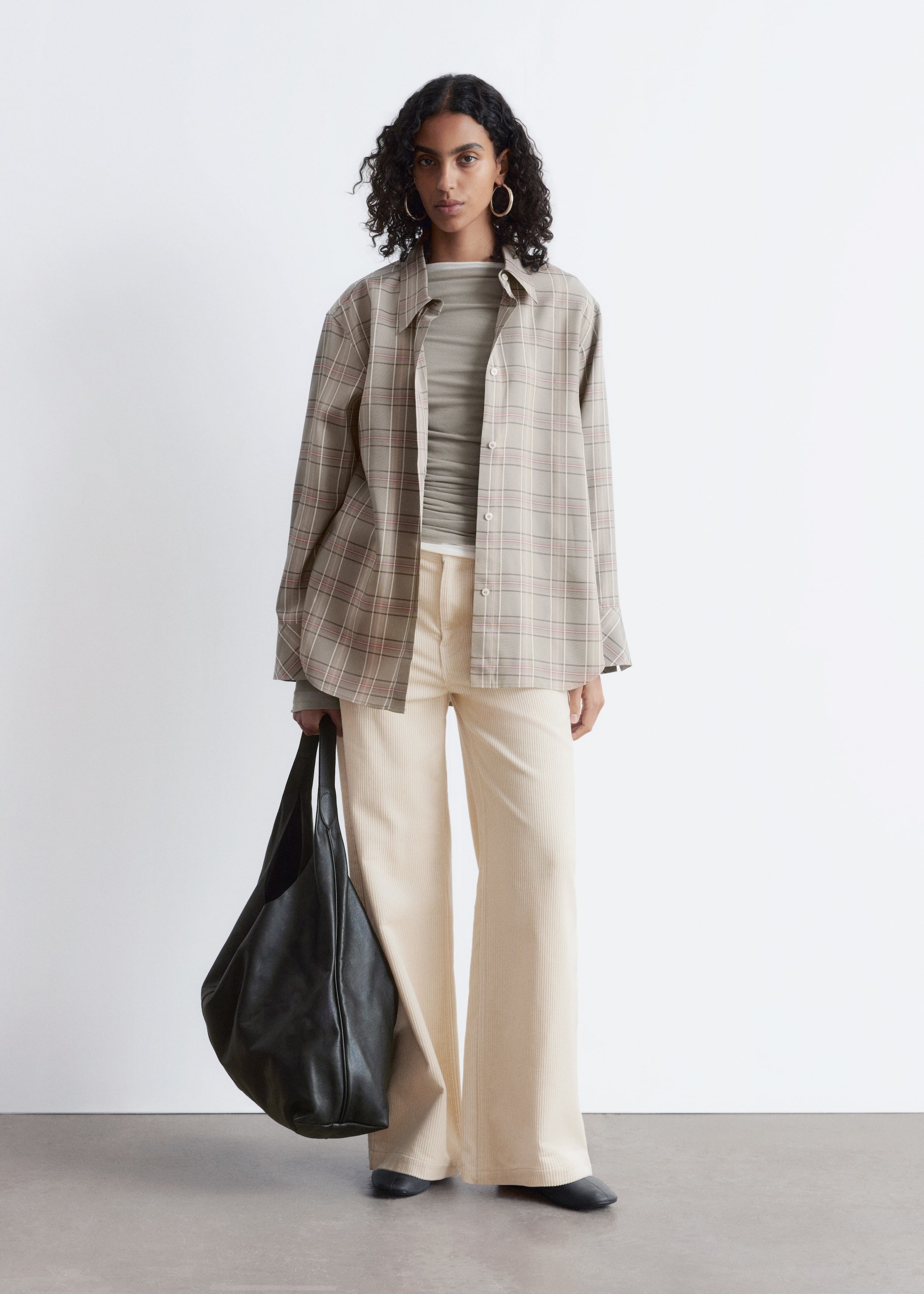 Visualizza immagine più grande: Una camicia oversize a quadri beige con bottoni, con sottili linee rosse e bianche, viene indossata aperta sopra un top a maniche lunghe a coste color oliva. Pantaloni a zampa di velluto a coste color crema completano l'outfit, insieme a una grande borsa morbida nera e scarpe basse nere, tutti presentati rivolti in avanti.