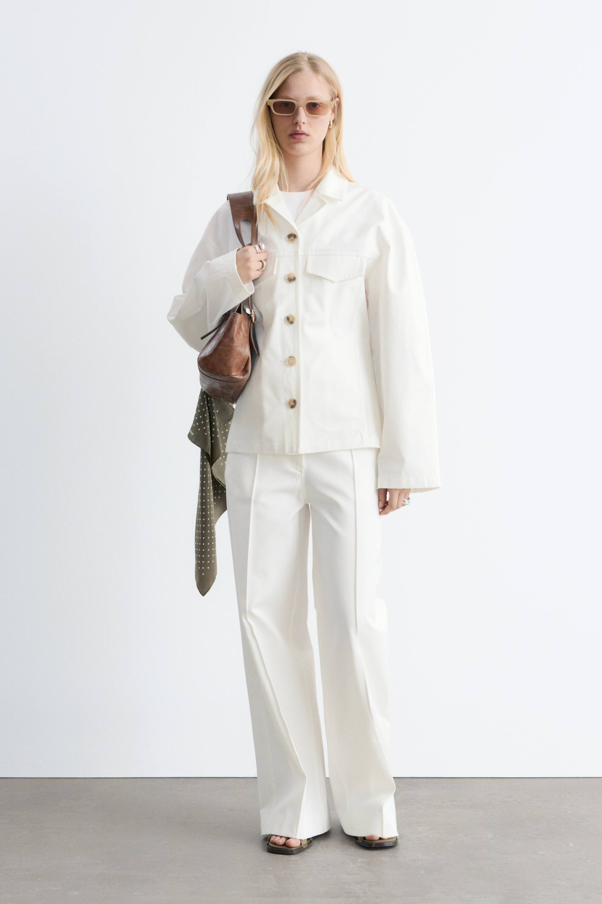 Vis større bilde: Modellen har på seg en hvit jakke i bomullstwill med enkelspent front, lysebrune knapper, hakkeslag og to brystlommer med klaff. Jakken er skåret i en slank silhuett med innsvinget midje. Modellen har også på seg matchende hvite bukser og bærer en brun skulderveske.