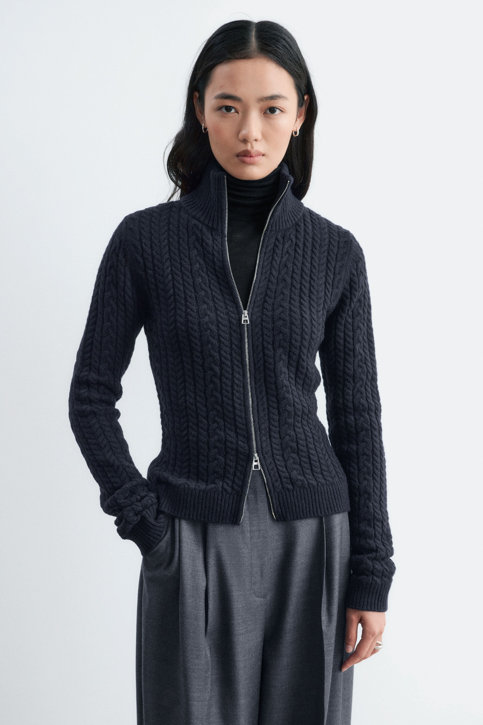 La modelo lleva un cárdigan de punto trenzado mixto en gris oscuro, superpuesto sobre un jersey de cuello alto negro y combinado con pantalones grises. El cárdigan de corte entallado presenta un cuello alto de doble capa acanalada y una cremallera frontal plateada de doble carro parcialmente abierta por ambos extremos. - 1