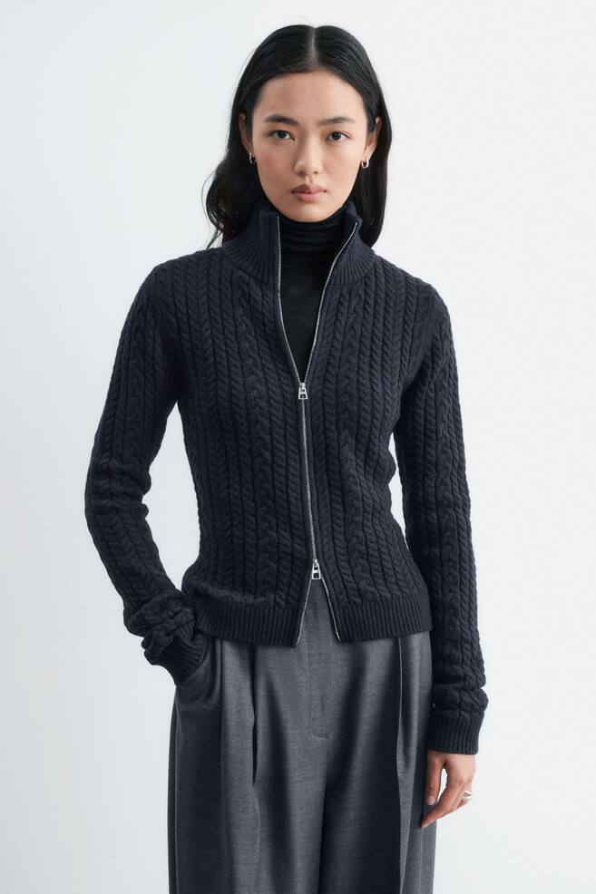 Het model draagt een donkergrijs, gemêleerd cable-knit vest, gelaagd over een zwarte coltrui en gecombineerd met een grijze broek. Het slim-fit vest heeft een hoge, dubbellaagse geribbelde kraag en een zilverkleurige two-way ritssluiting vooraan die aan beide uiteinden gedeeltelijk open is. - 1