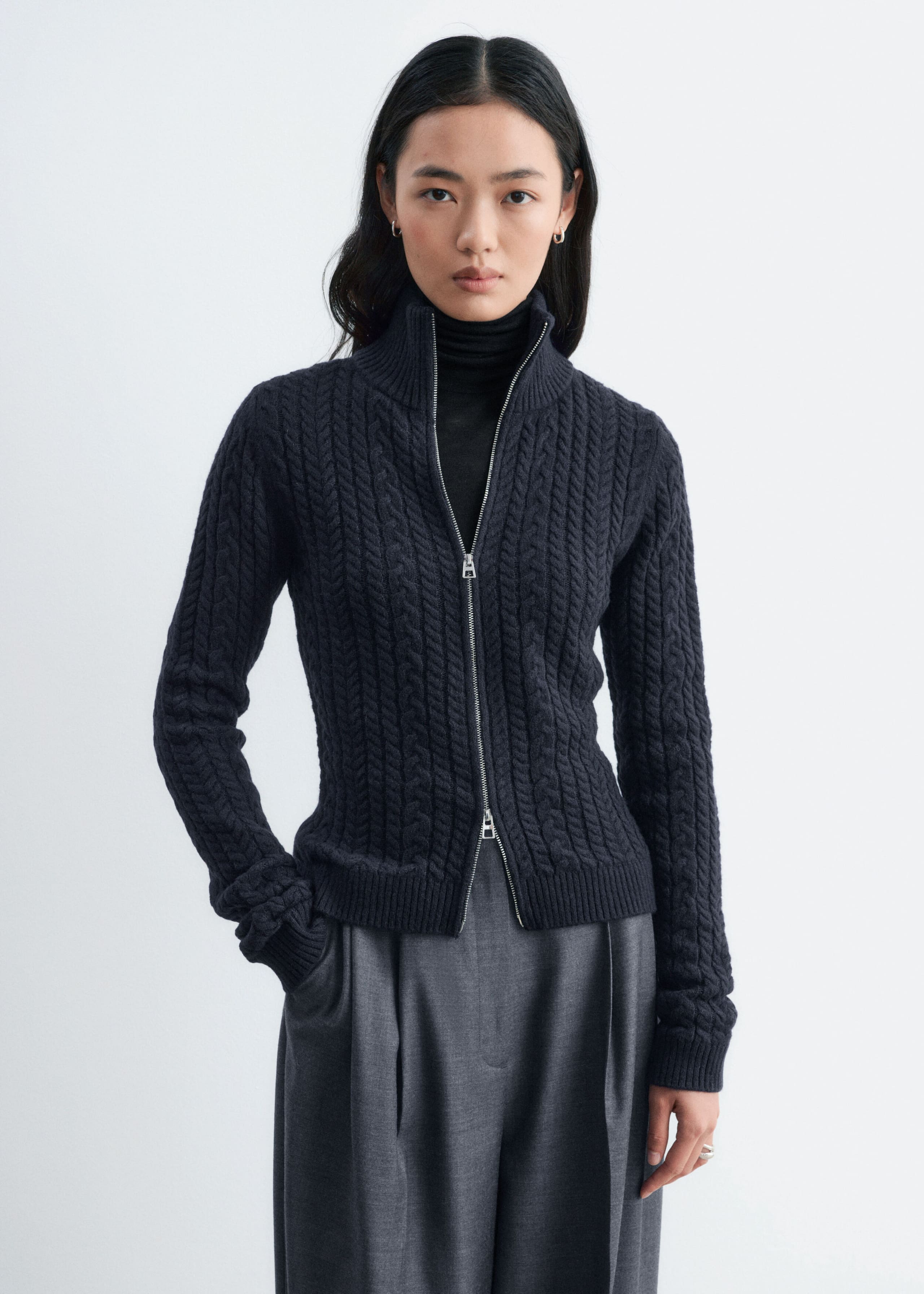 A dark grey mixed cable-knit cardigan is being worn by the model, layered over a black roll-neck and paired with grey trousers. The slim-fit cardigan features a high, double-layer ribbed collar and a silver two-way front zip that is partially open at both ends.