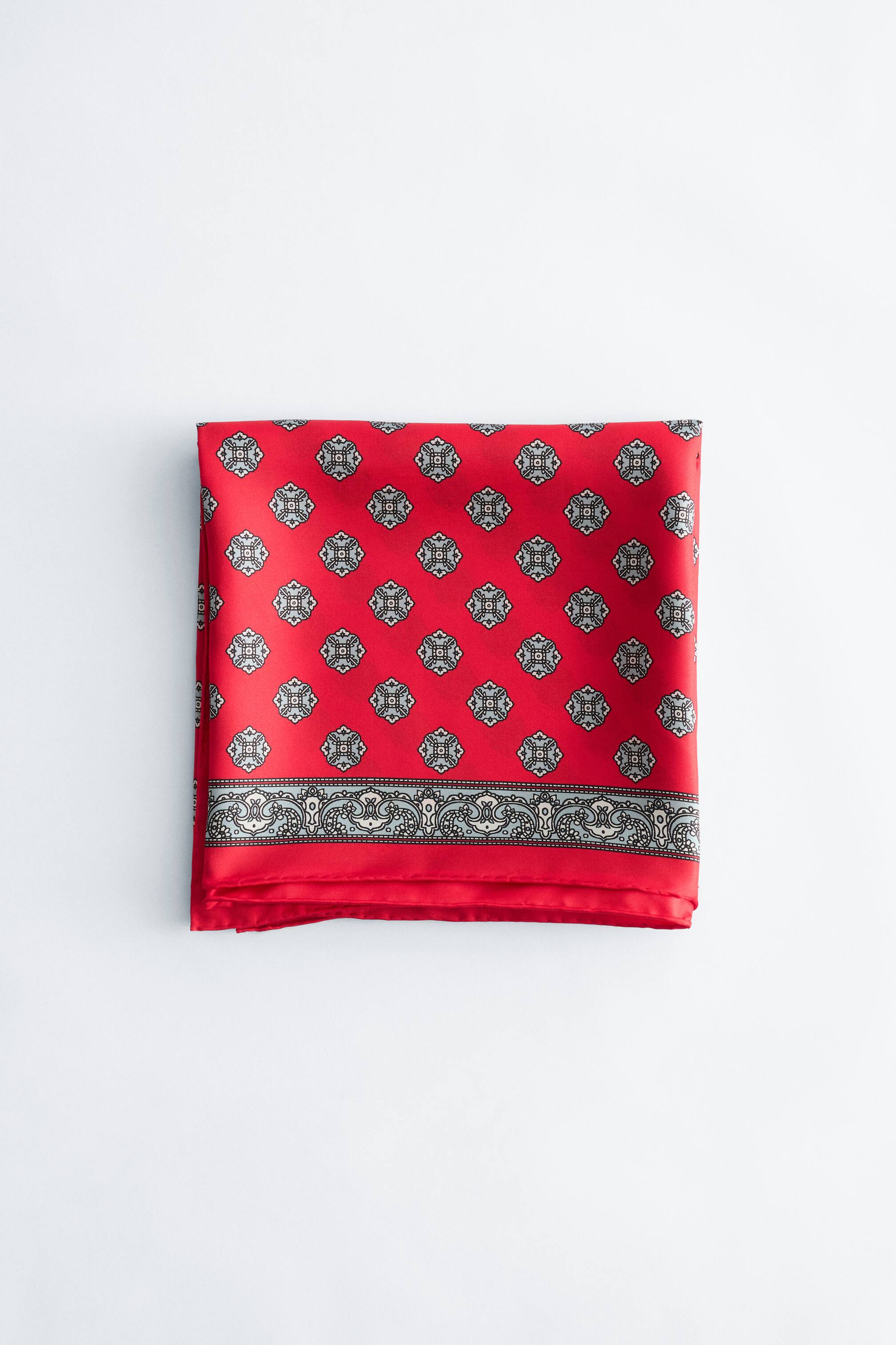 View larger image: A red silk scarf displays a repeating pattern of black, white and grey stylised quatrefoil motifs. A wide decorative border featuring intricate scrolling runs along its lower edge.