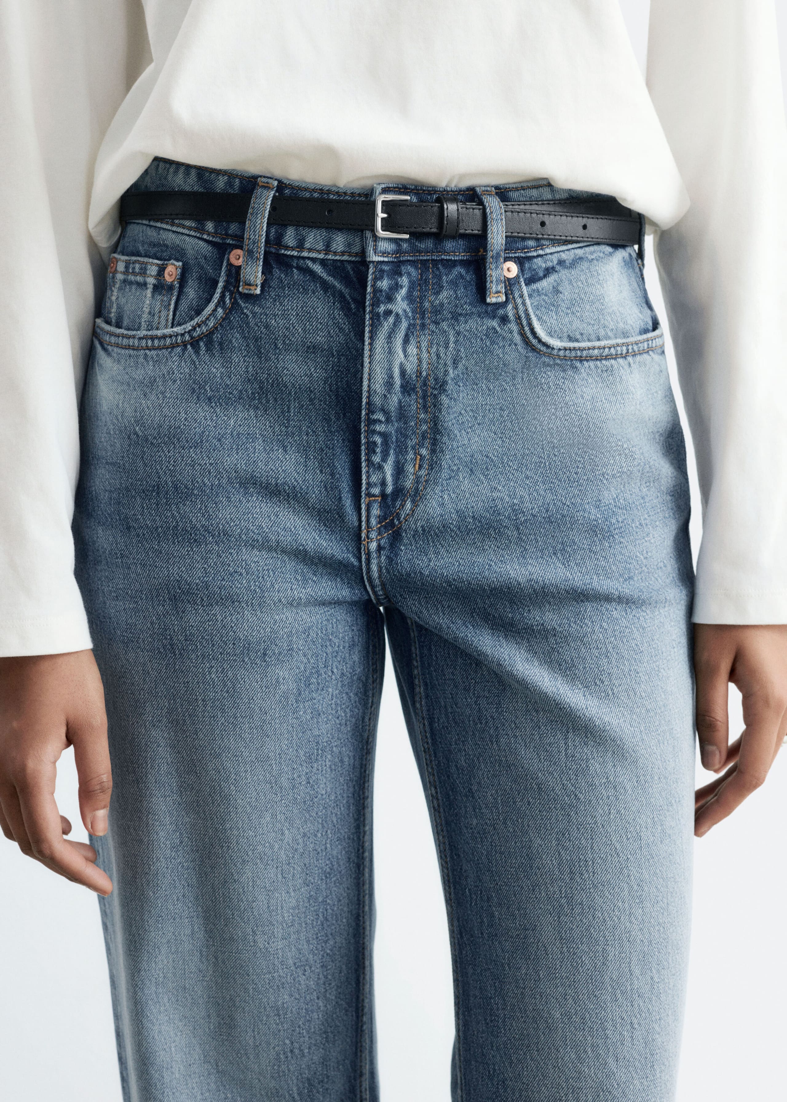 The model is wearing blue mid-rise, straight-leg jeans, styled with a thin black belt and a tucked-in white long-sleeve top. The jeans feature a classic five-pocket design with copper-toned rivets.
