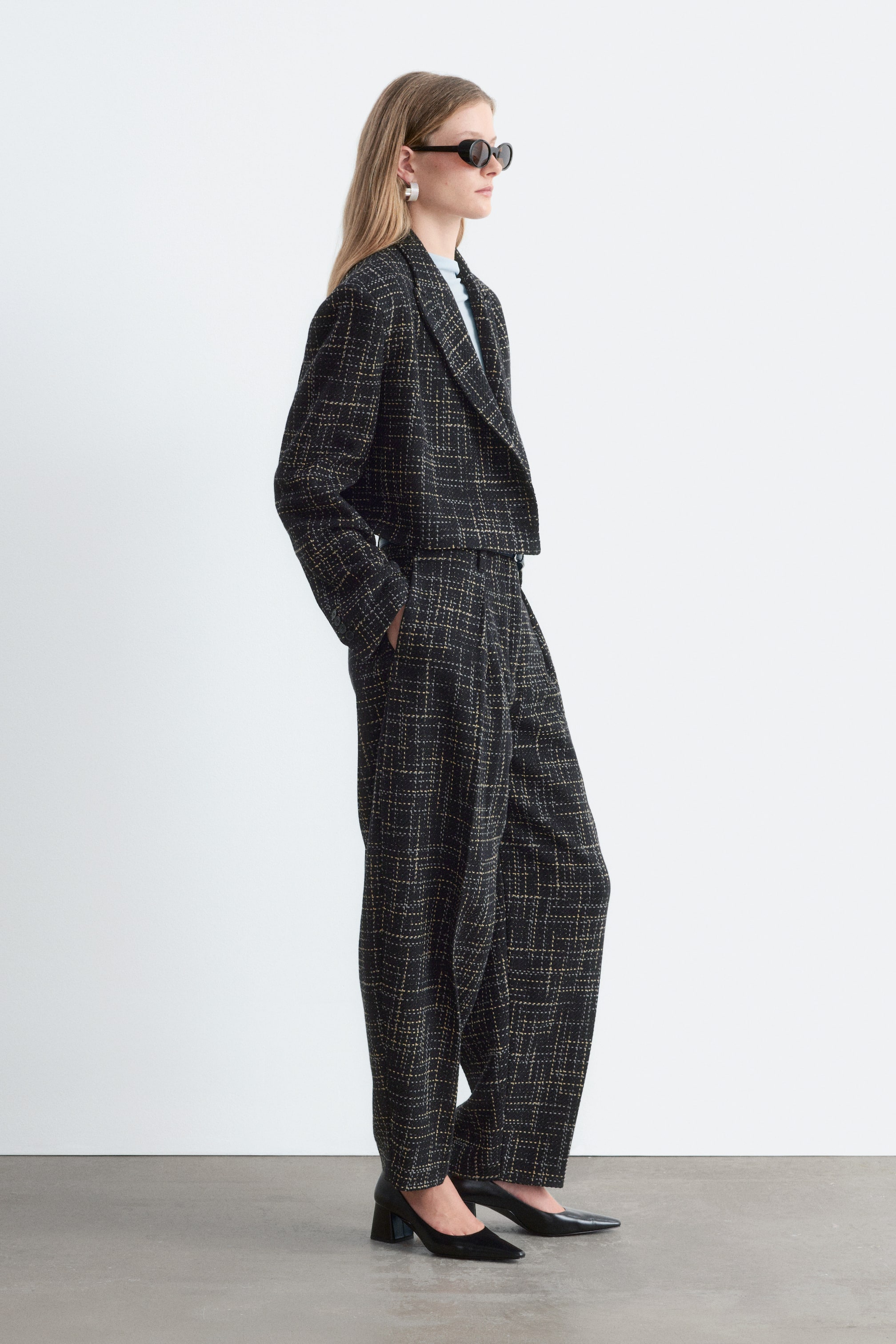 Visa större bild: En kort blazer i ett svart- och gult texturerat rutmönster visas på en modell som vänder sig åt höger, vilket framhäver de markerade axlarna, de klassiska slaglappen och den rent avslutade fållen. Blazern är lagd över en ljusblå topp och matchad med vida byxor i samma stil för att visa dess korta, strukturerade siluett.