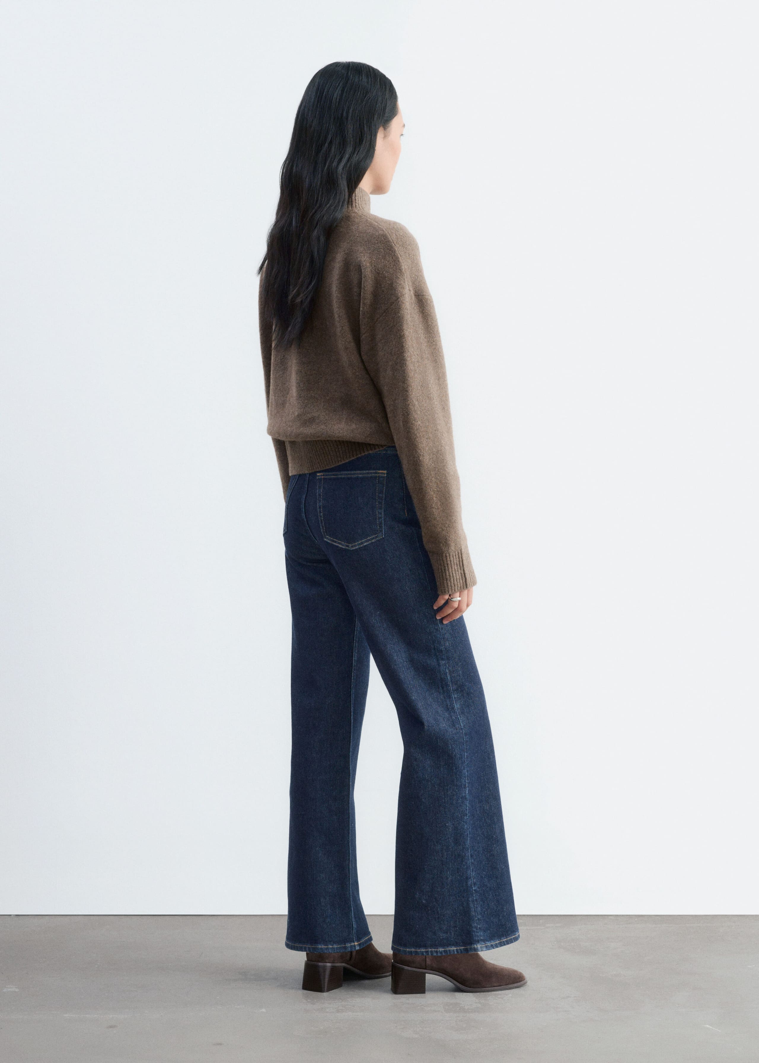 Het model draagt een bruine gebreide trui met lange mouwen, met een geribde col, verlaagde schoudernaden en een geribde zoom. De losvallende trui is gecombineerd met donkerblauwe wide-leg jeans en bruine laarzen met hak.