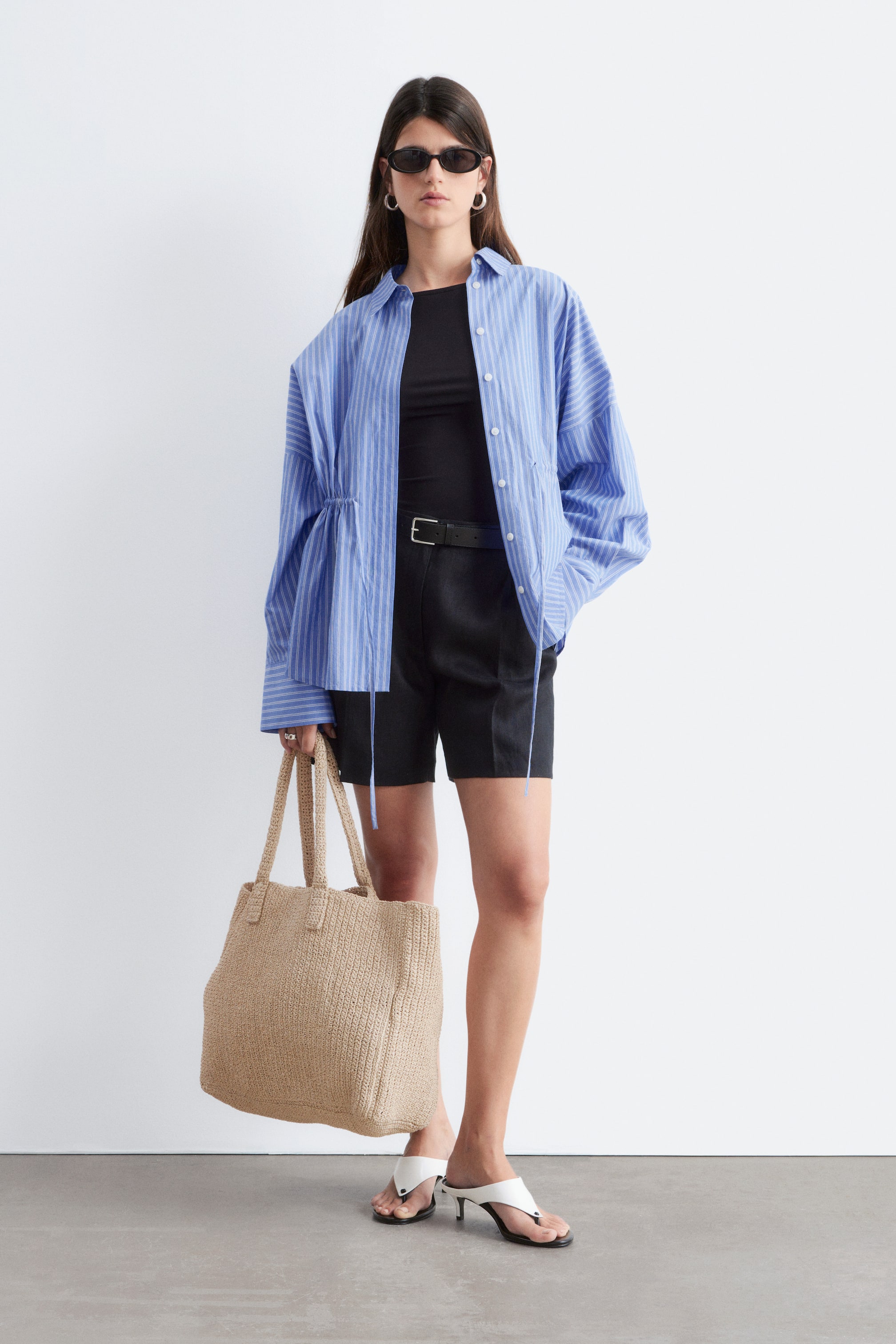 Größeres Bild ansehen: Ein blau-weiß gestreiftes Button-up-Hemd wird offen über einem schwarzen Oberteil getragen, mit einem klassischen Kragen, langen Ärmeln und Kordelzugdetails, die die Seiten raffen.