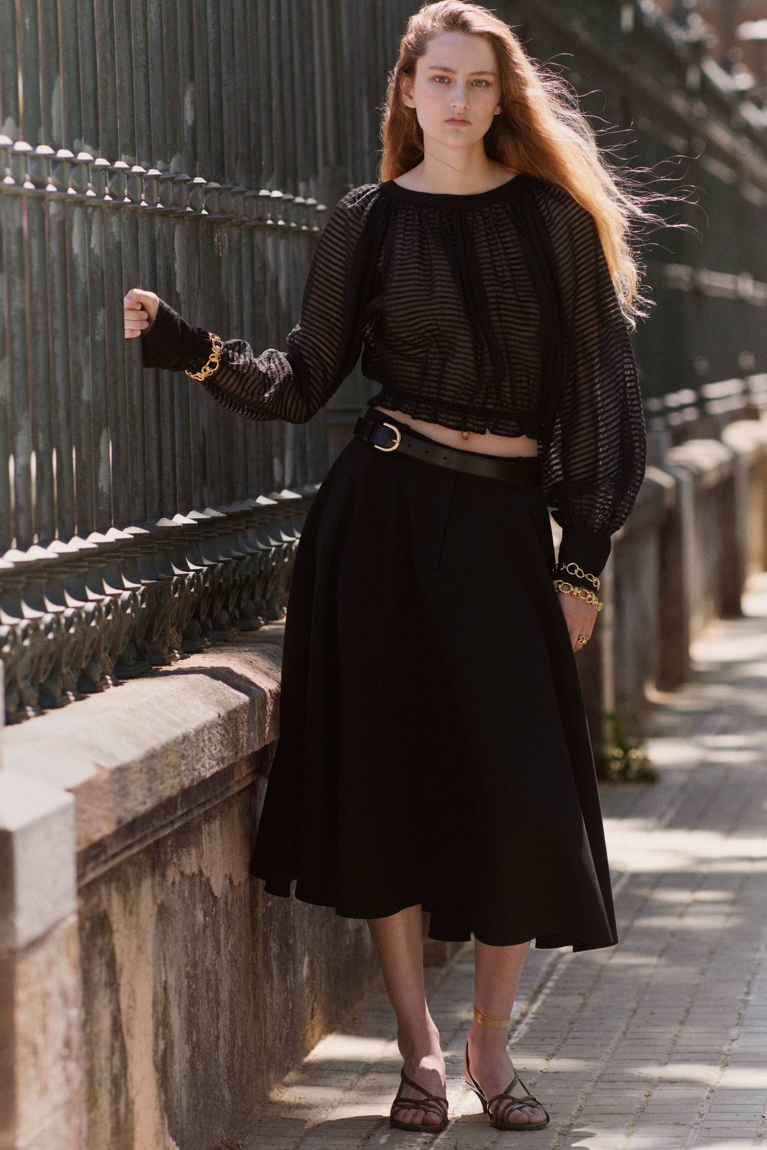 A black blouse is facing front, featuring a sheer fabric with a subtle horizontal striped texture. It is designed with a gathered round neckline, long balloon sleeves with gathered cuffs, and a cropped, elasticated hemline with a delicate ruffle. - 1