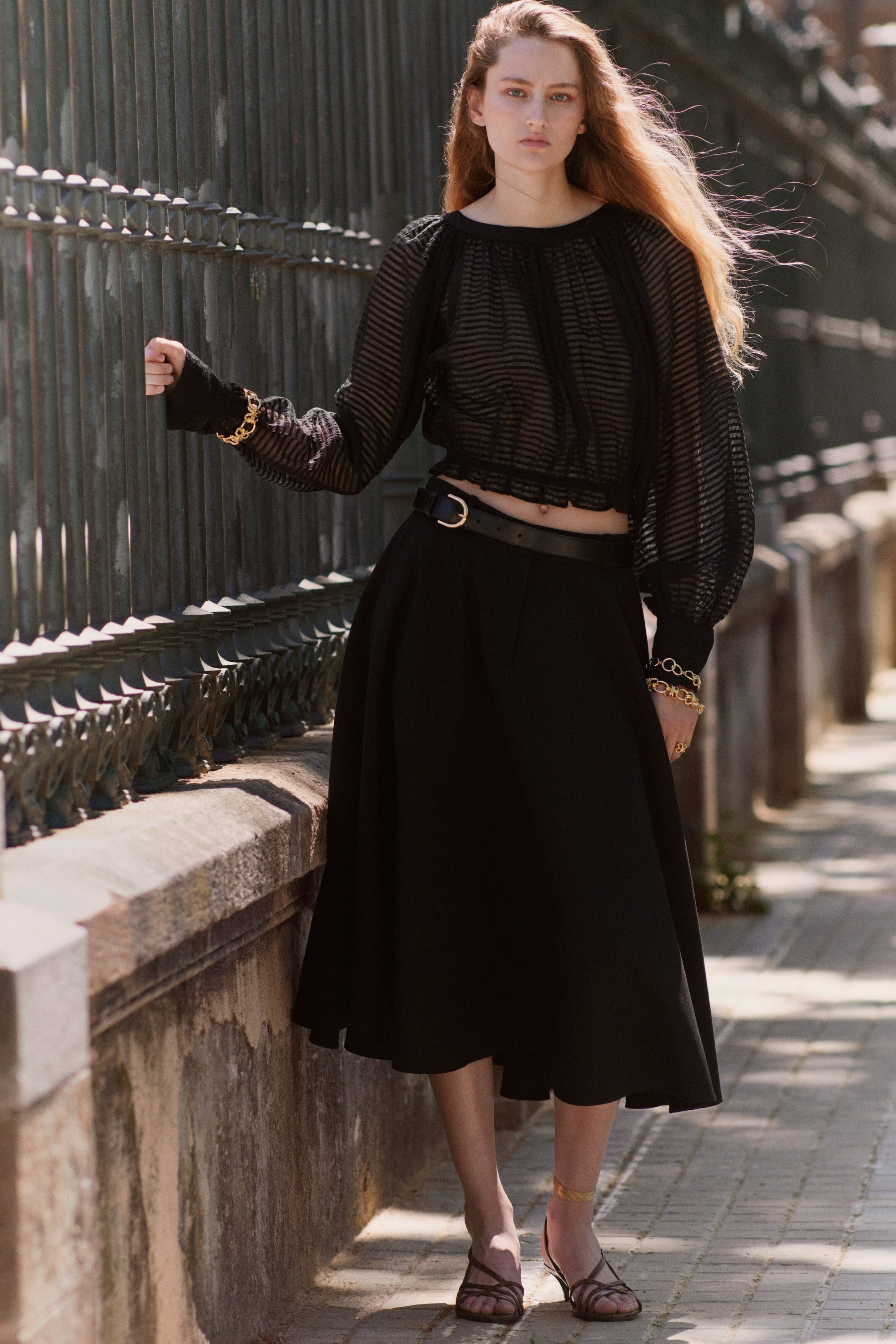 View larger image: A black sheer top with horizontal stripes, a gathered round neckline, voluminous long sleeves, and a cropped elasticated hem is styled with a black midi A-line skirt, cinched at the waist with a black belt featuring a gold-tone buckle.
