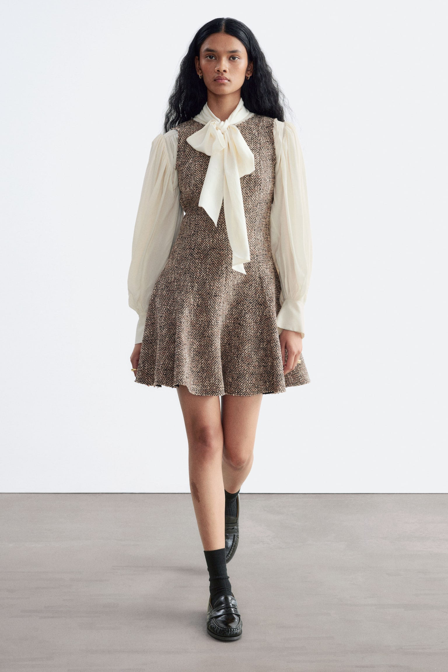 O rochie mini cu margini nerelucrate, cu model herringbone din tweed maro, este purtatÄ de model. Rochia fÄrÄ mâneci are o fustÄ evazatÄ Èi este suprapusÄ peste o bluzÄ crem, cu mâneci lungi, cu o fundÄ proeminentÄ la gât. - 1