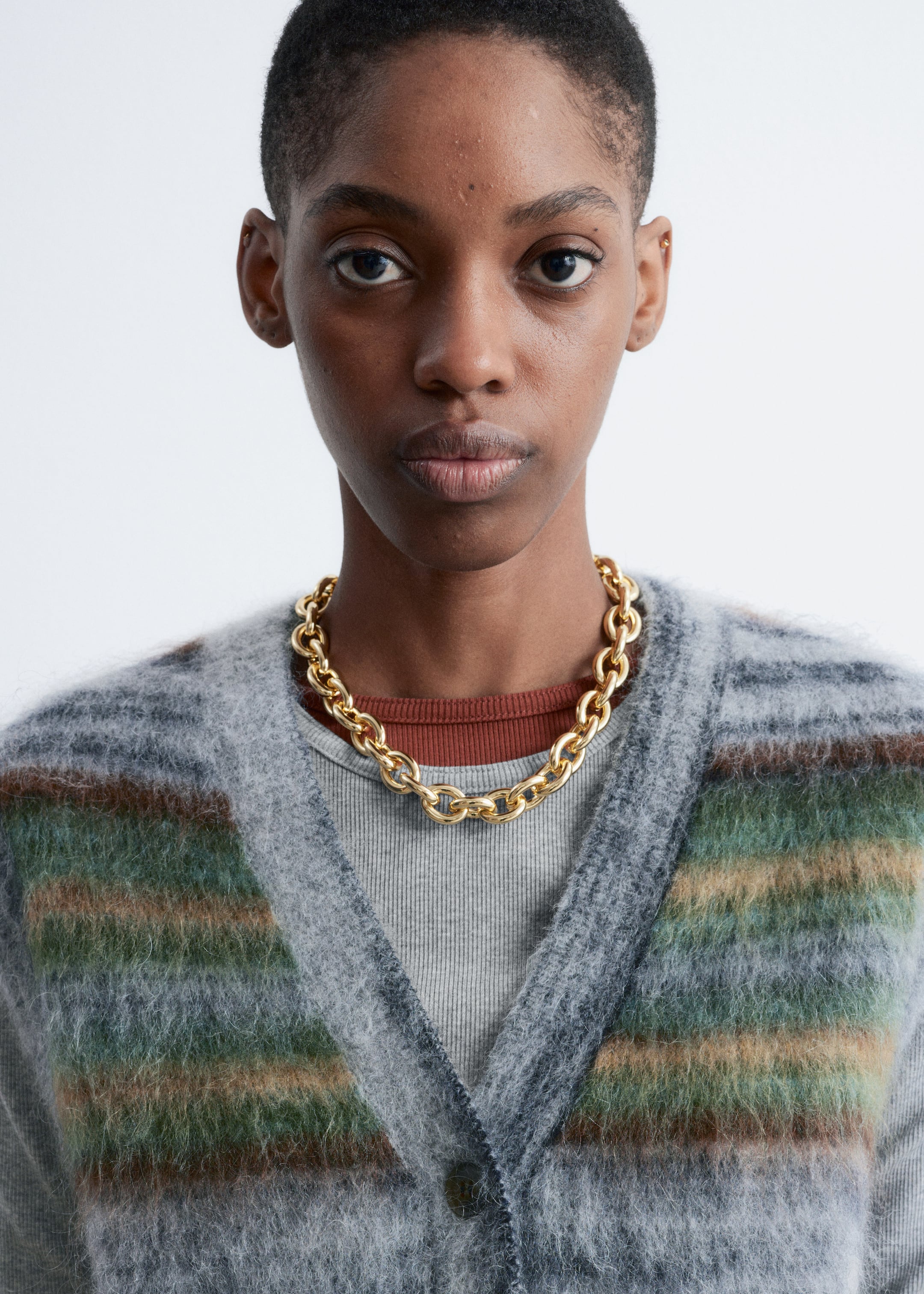 Visualizza immagine più grande: Un cardigan fuzzy con scollo a V, caratterizzato da righe orizzontali in grigio, verde e marrone, viene presentato da un'angolazione frontale. È stratificato sopra una t-shirt grigia a coste, indossata sopra una t-shirt con collo a giro di colore ruggine. Una robusta collana a catena dorata riposa sui colli stratificati.