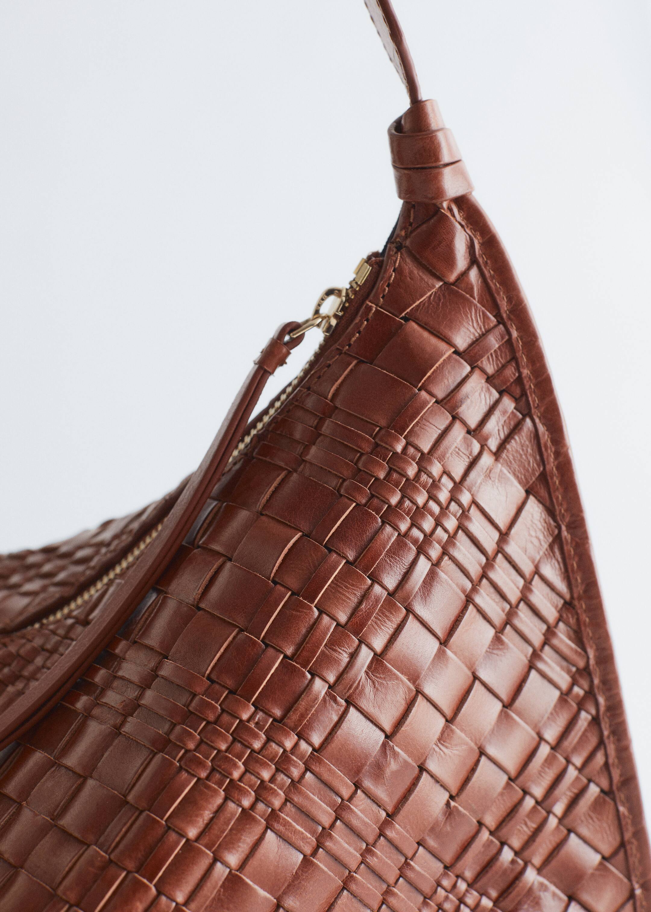 Größeres Bild anzeigen: Eine braune Ledertasche wird aus einem schrägen, nahen Blickwinkel gezeigt, wobei ihre komplizierte gewebte Textur hervorgehoben wird. Sie verfügt über einen geschwungenen oberen Rand mit einem messingfarbenen Reißverschluss und einem langen Lederzug, alles aus verflochtenen Lederstreifen gefertigt.