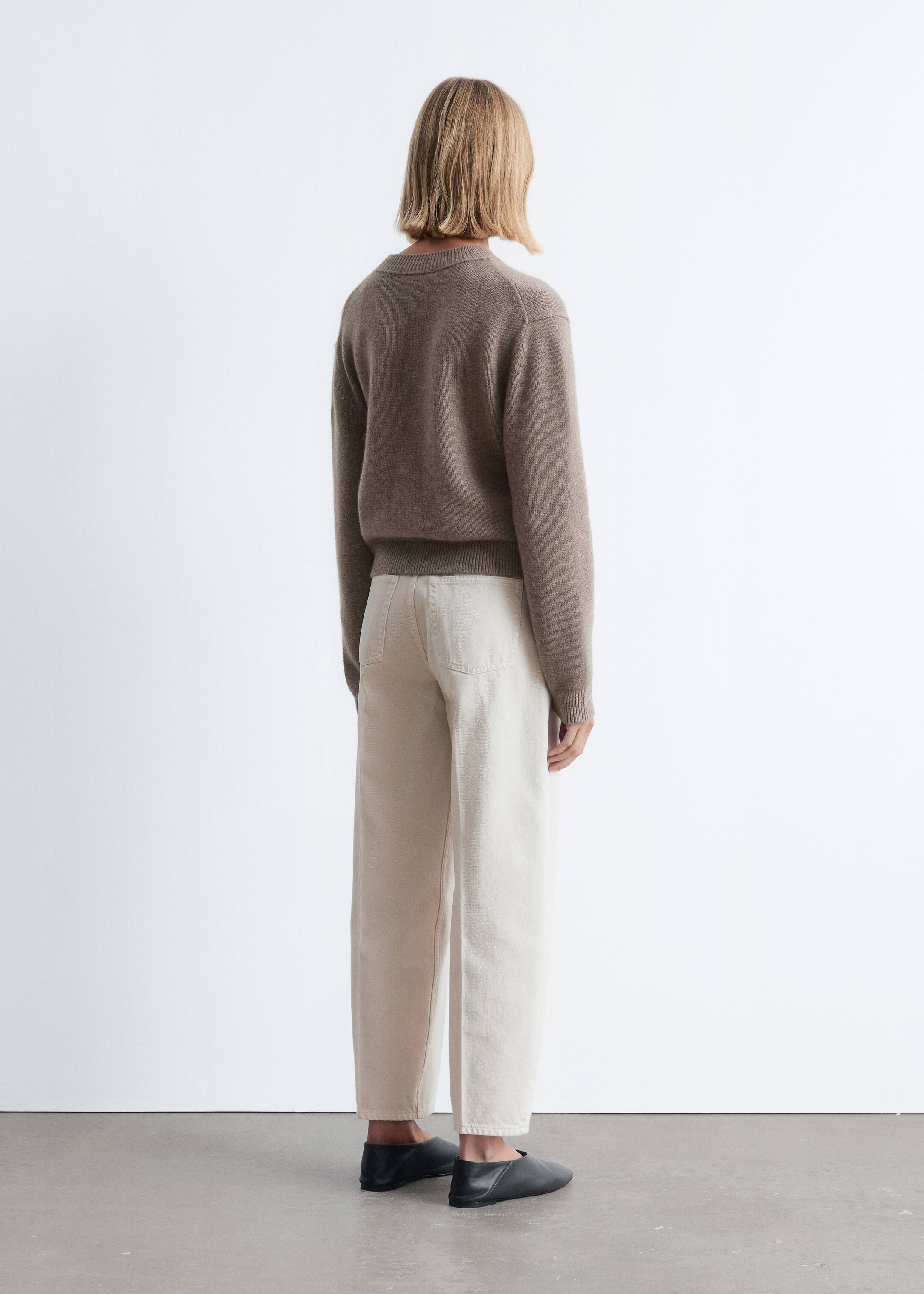Ingrandisci l'immagine: Un maglione corto color talpa con finiture a coste e spalle scese è indossato da una modella, che sta in piedi con la schiena rivolta verso l'osservatore. Il maglione è abbinato a pantaloni a gamba larga di colore chiaro e scarpe basse scure.