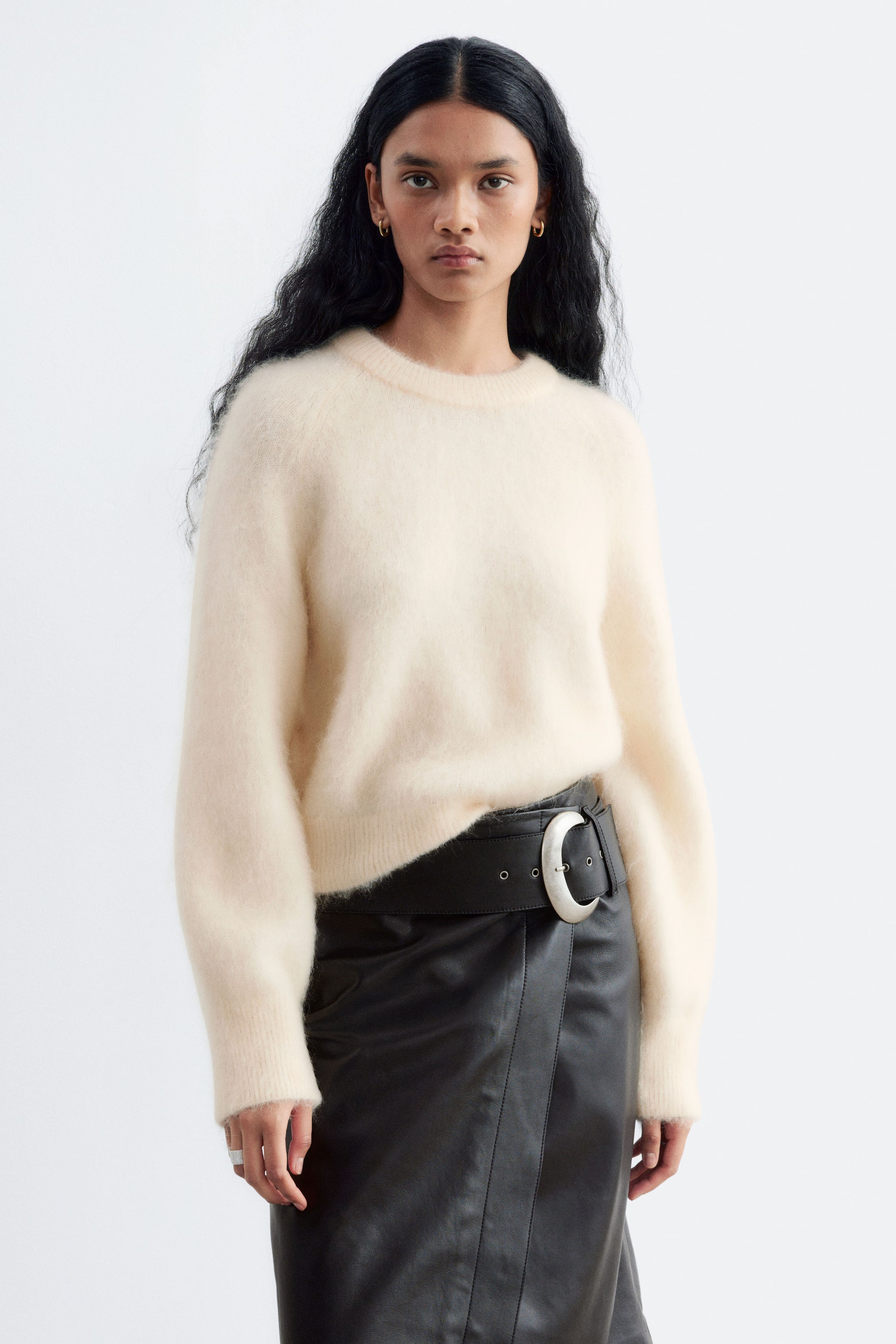 Agrandir l'image: Un pull en maille duveteuse de couleur crème est porté par le mannequin, avec un col rond côtelé et de longues manches raglan à larges poignets. Le pull est partiellement rentré à la taille dans une jupe en cuir noire avec une large ceinture.