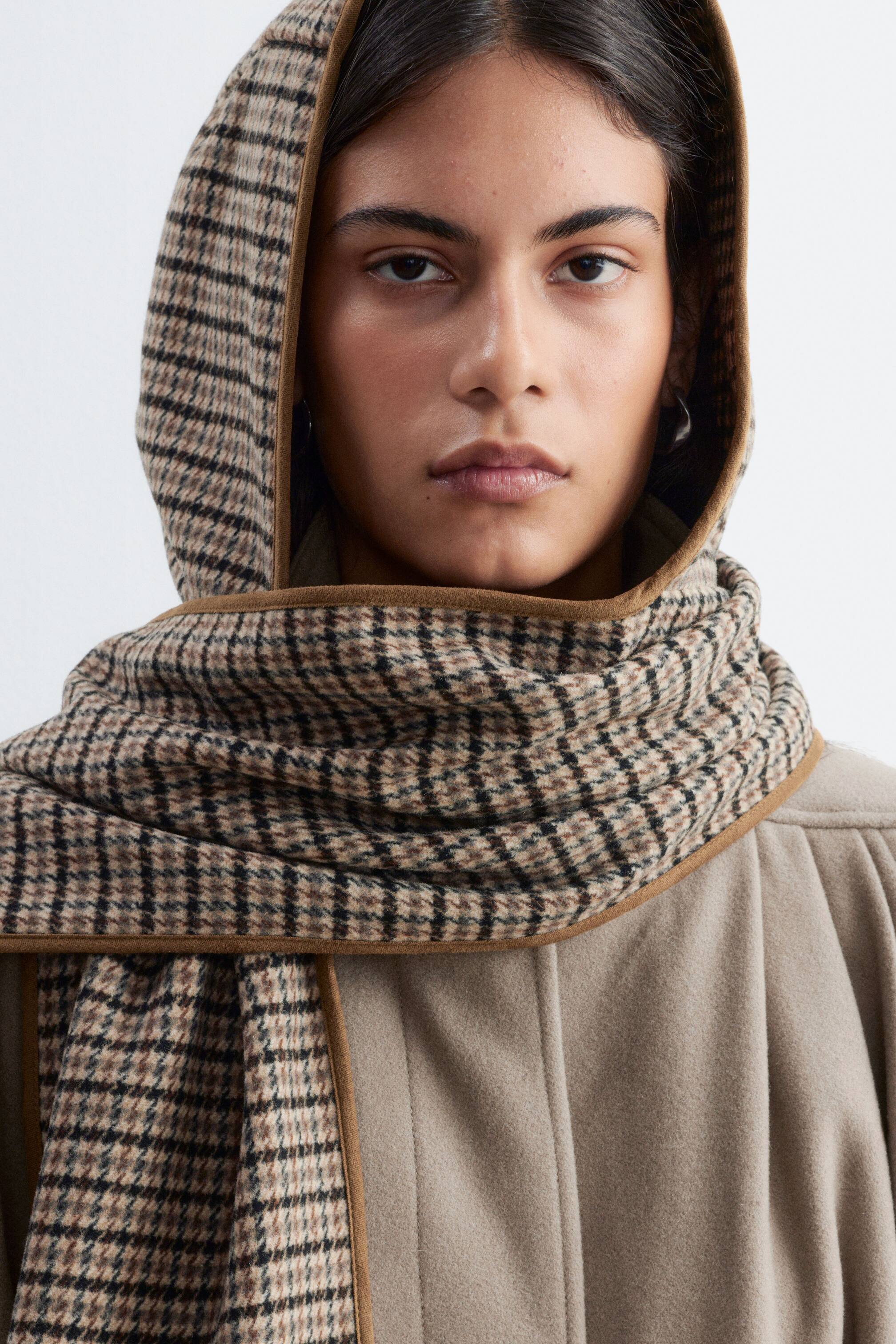 Agrandir l'image: Une capuche à motif pied-de-poule avec une écharpe intégrée est portée par le mannequin, couvrant la tête et enveloppant le cou. Cet accessoire en mélange de laine présente un biais brun sur les bords et se superpose à un manteau beige.