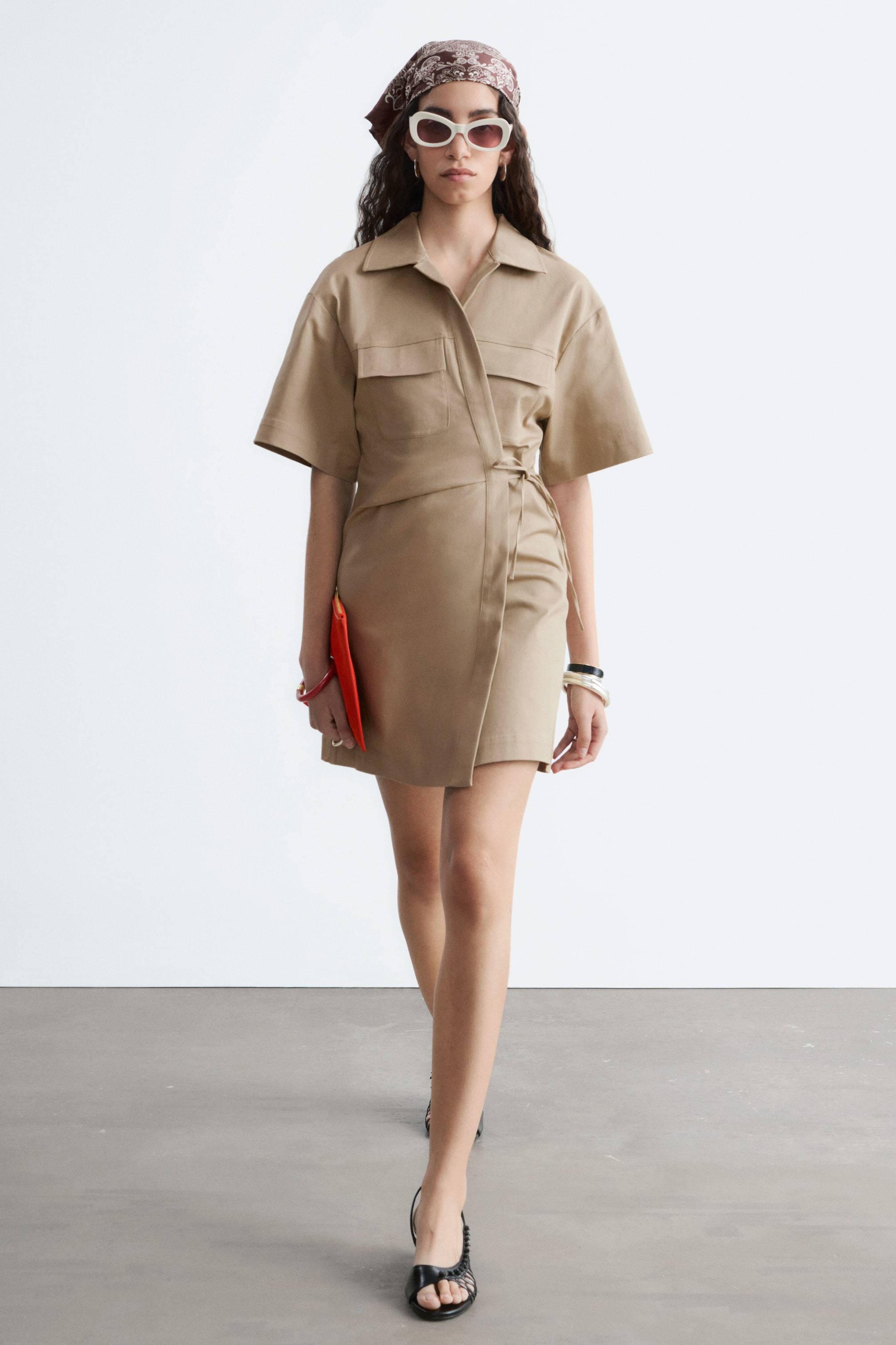 Visa större bild: En beige miniklänning med öppen krage, korta vida ärmar och två bröstfickor med lock bärs av modellen. Klänningen är designad med en omlott-silhuett och ett knytband i midjan. Modellen bär även en orange kuvertväska och svarta sandaler.