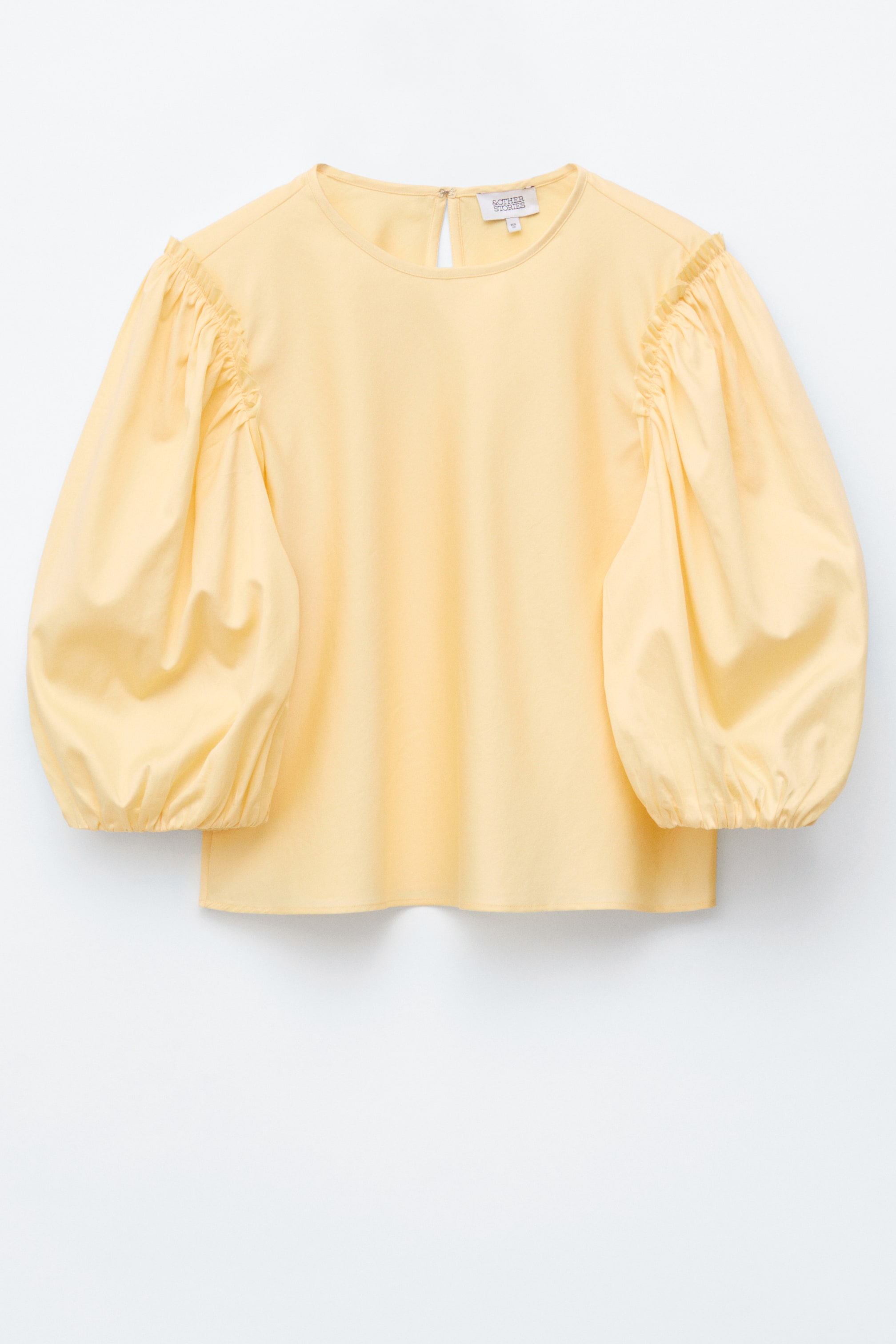Grotere afbeelding bekijken: Een lichtgele blouse wordt frontaal gepresenteerd, met een ronde halslijn, volumineuze ballonmouwen die bij de schouders zijn geplooid en elastische manchetten, en een kleine sleutelgat sluiting aan de achterkant van de hals.
