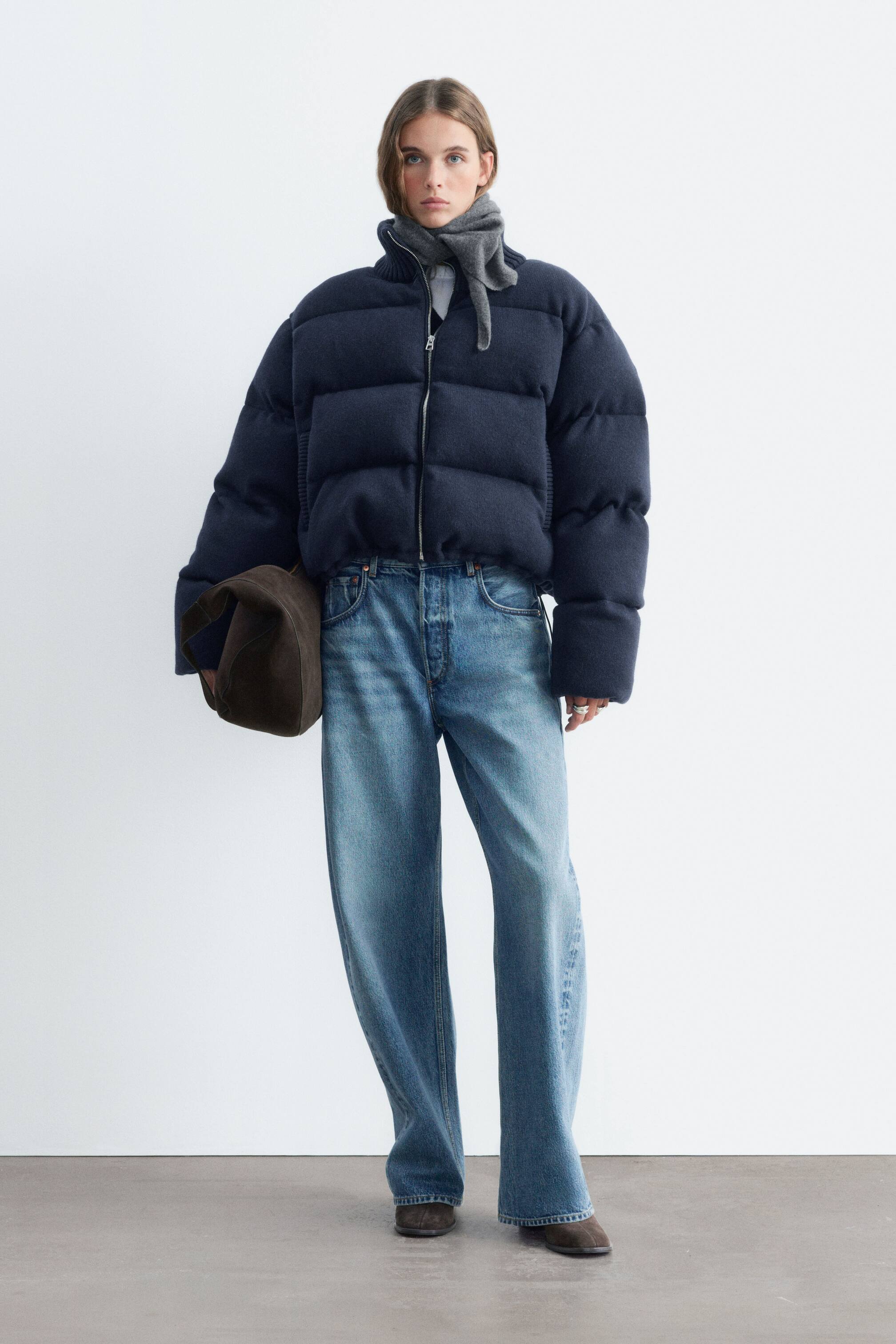 View larger image: A navy blue knitted puffer jacket with horizontal quilting and a high collar is being worn by the model. The jacket is zipped up and styled with a grey scarf, wide-leg blue jeans, and a dark brown bag held in one hand.