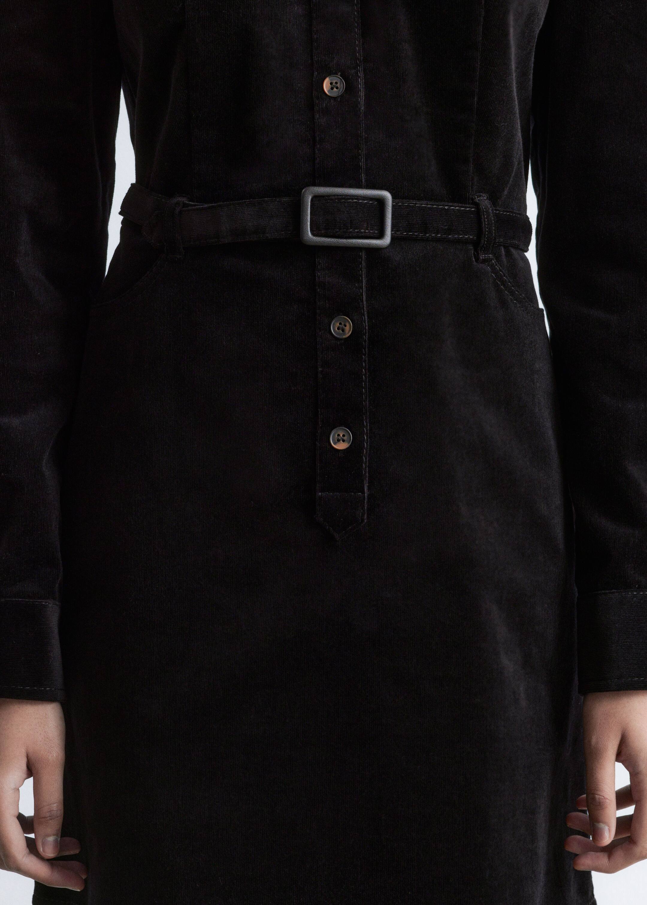 Agrandir l'image: Une robe en velours côtelé noir est présentée de face, mettant en valeur un col de style chemise, une patte de boutonnage avant avec des boutons marron, et une ceinture en velours côtelé assortie qui cintre la taille avec une boucle rectangulaire noire. Des poches latérales en biais sont également visibles.