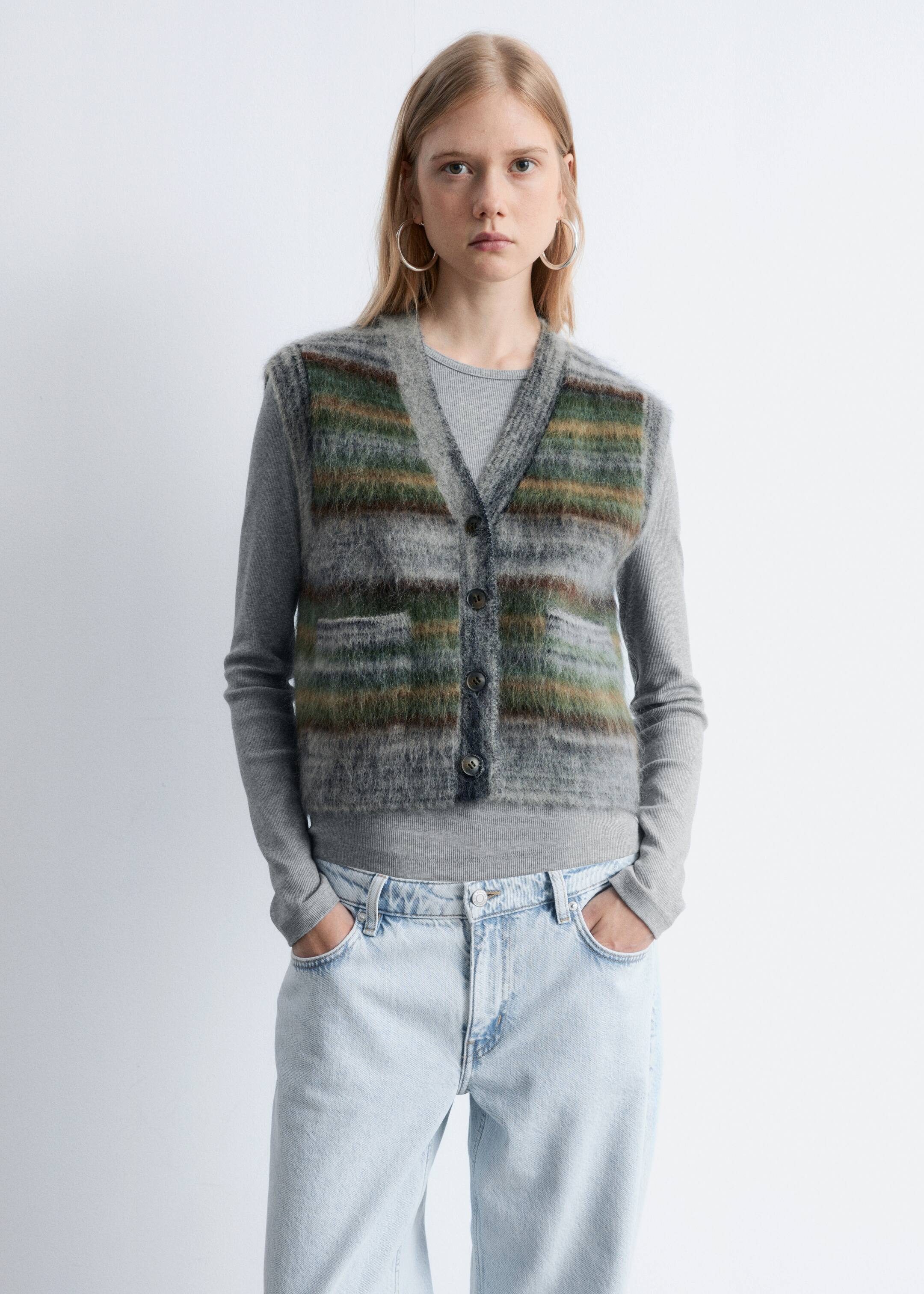Visualizza immagine più grande: Un gilet cardigan con scollo a V sfocato viene presentato frontalmente, mostrando strisce orizzontali in tonalità di grigio, verde e marrone. Presenta una chiusura frontale a quattro bottoni e due tasche a toppa, ed è stratificato sopra una maglia a maniche lunghe grigia e jeans a lavaggio chiaro.