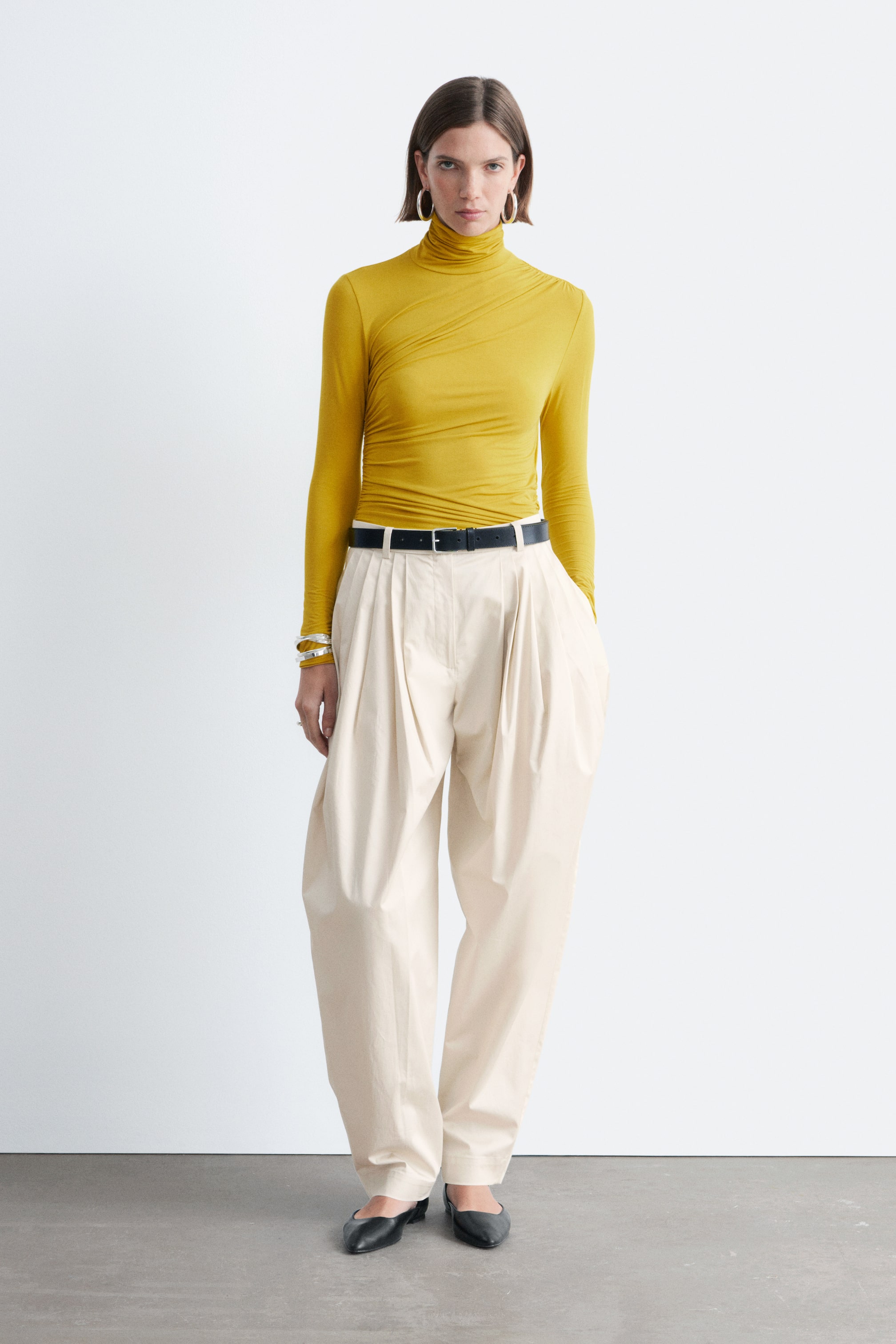 Ingrandisci l'immagine: Un top a collo alto giallo senape, caratterizzato da un tessuto fluido e cuciture raccolte che creano un effetto drappeggiato, è indossato dalla modella. Ha maniche extra lunghe e una vestibilità aderente, ed è infilato in pantaloni di colore chiaro.