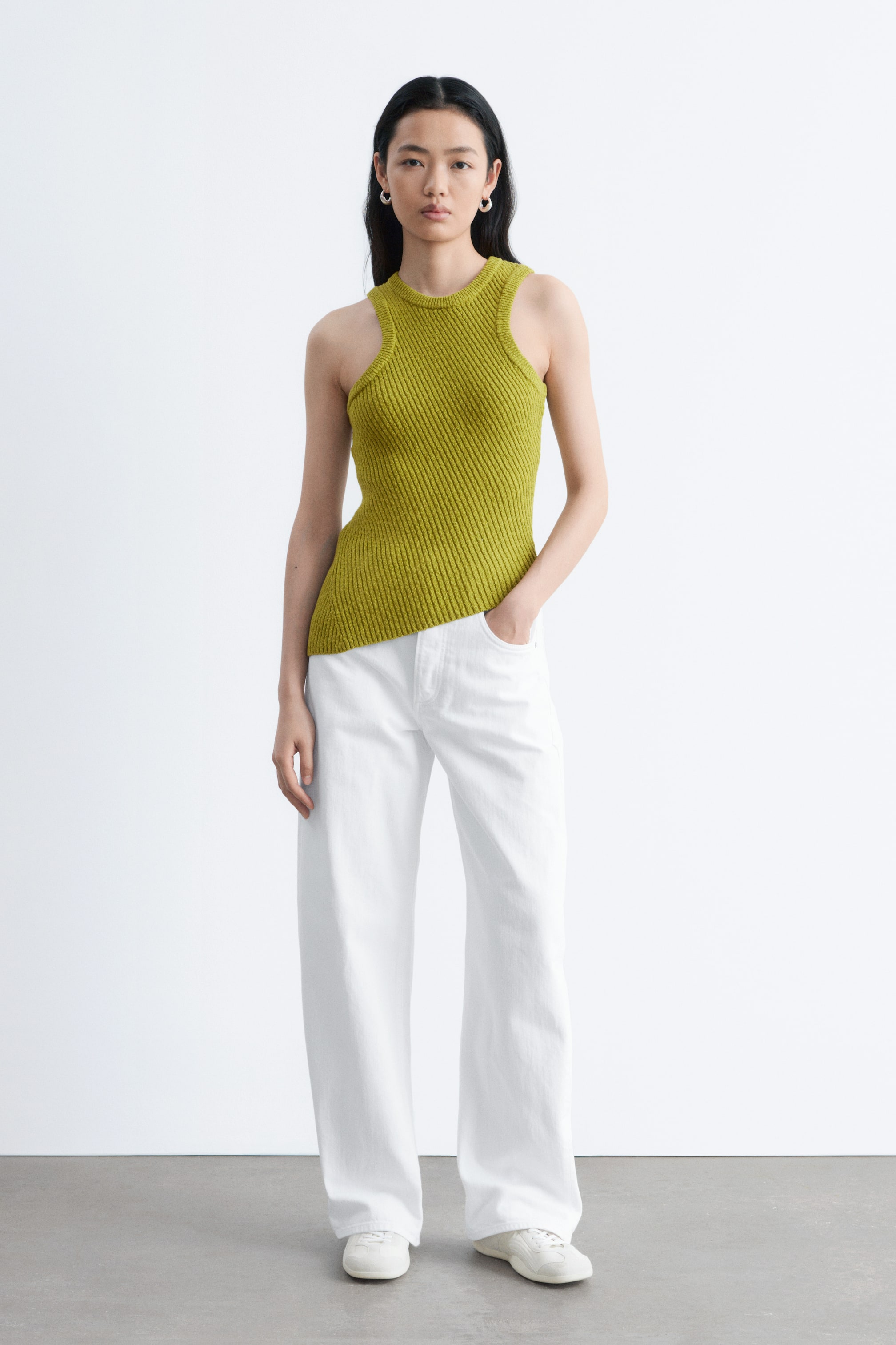 Ver imagen más grande: Un top de punto acanalado verde lima presenta un cuello redondo y sisas recortadas. Muestra un patrón acanalado diagonal y un dobladillo asimétrico, y se combina con pantalones blancos de pierna ancha.
