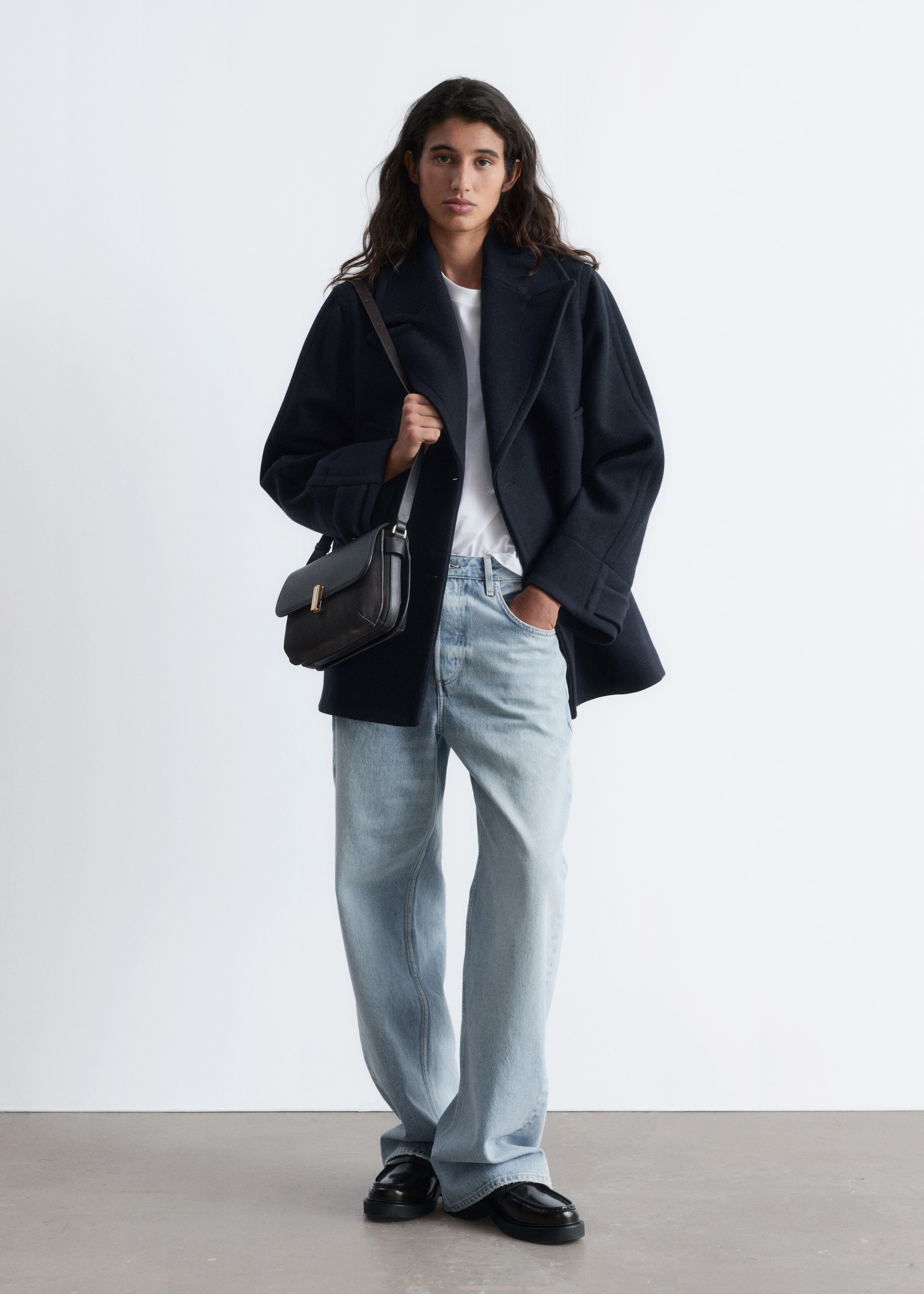 View larger image: A full outfit is being presented front-facing, consisting of a dark navy double-breasted peacoat with wide notch lapels, layered over a white crew-neck t-shirt and light wash, relaxed-fit wide-leg jeans. Black chunky loafers are being worn, and a black structured leather shoulder bag with a gold-toned clasp is being carried.