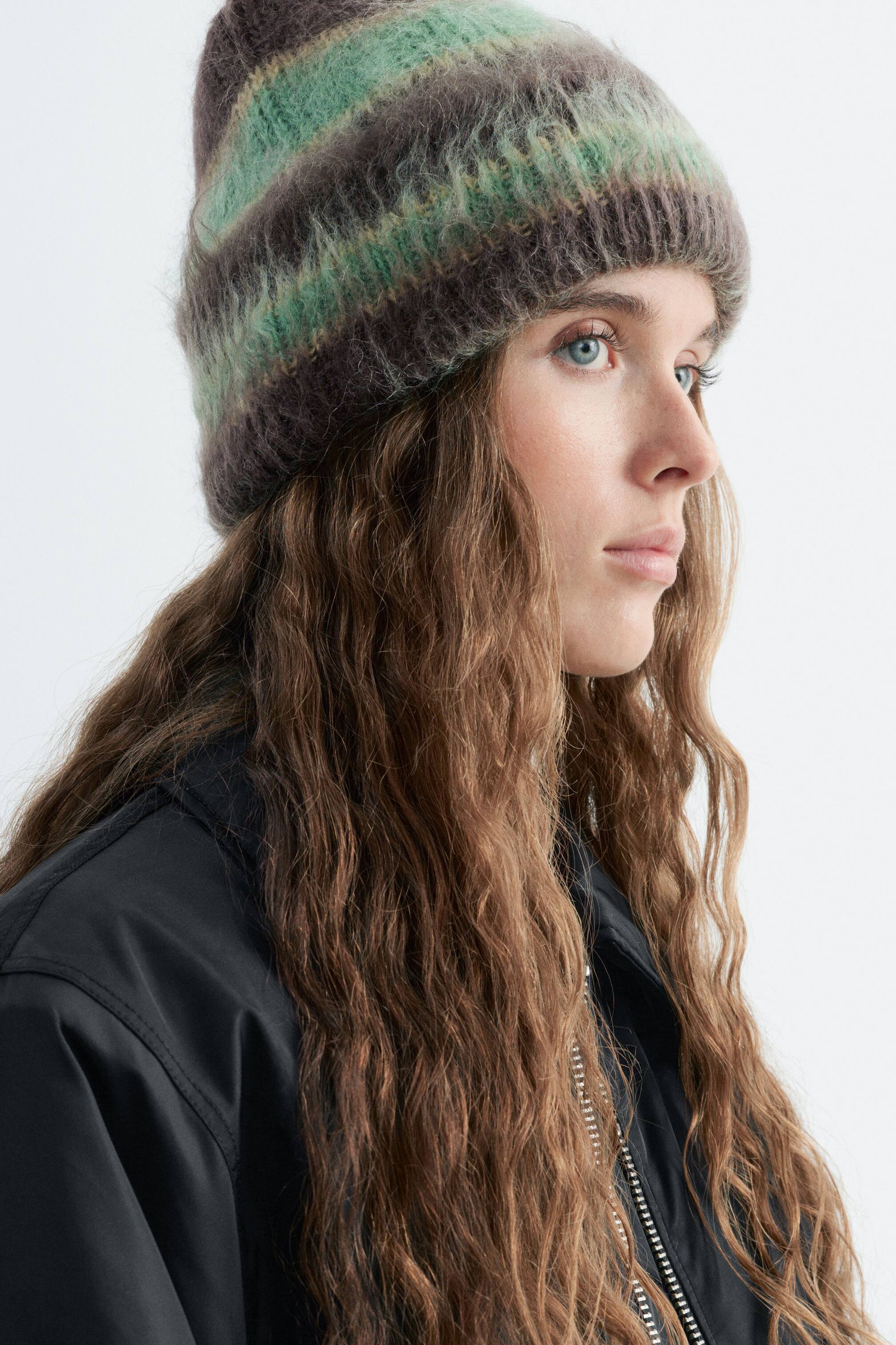 Visa större bild: En mjuk, texturerad stickad beanie med horisontella ränder i grönt och mörkbrunt bärs av modellen. Den enkelvikta beanie-hatten har en ribbad kant och matchas med en svart jacka med krage.