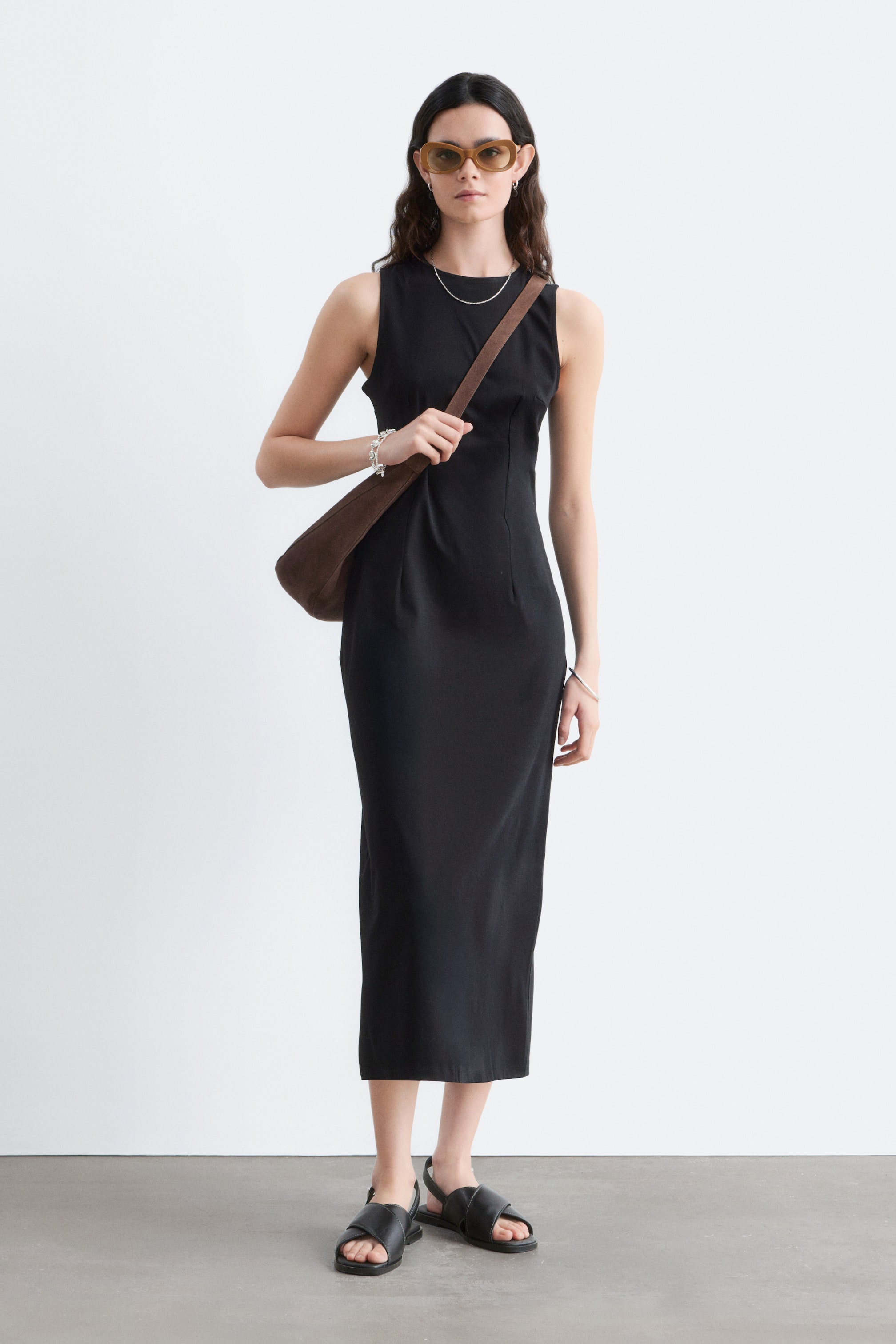 Agrandir l'image: La robe midi noire sans manches présente un col rond et une coupe ajustée qui épouse les formes du corps, s'étendant jusqu'à une longueur midi. Elle affiche des coutures verticales sur mesure à l'avant, avec une bandoulière marron traversant la poitrine et le sac reposant sur la hanche.