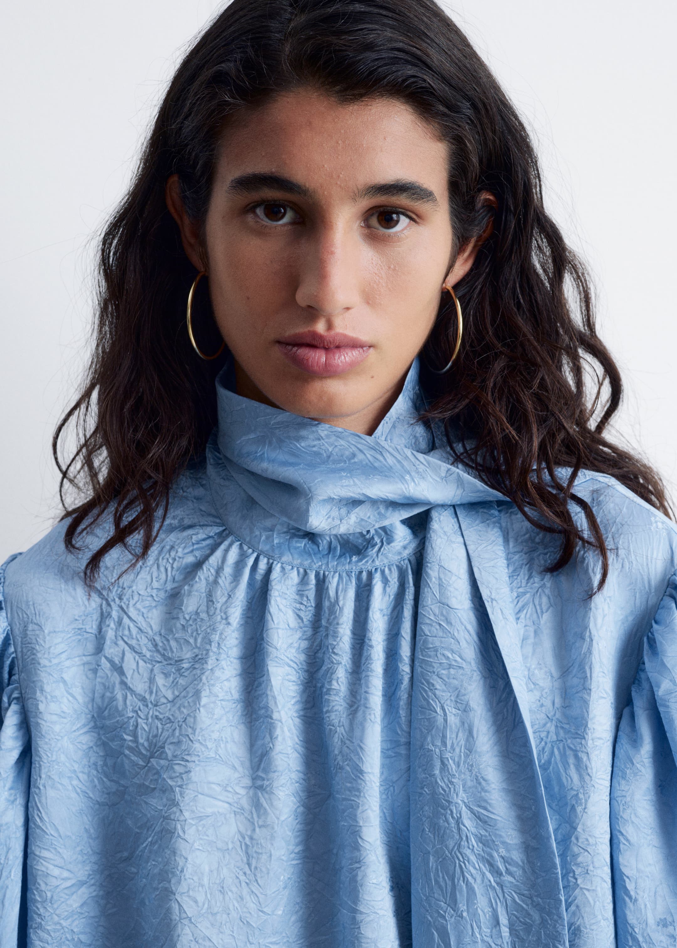 Grotere afbeelding bekijken: Een lichtblauwe blouse heeft een gekreukelde textuur door de hele stof. Het is ontworpen met een gedrapeerde hoge halslijn die overgaat in een geïntegreerd strikdetail en toont geplooide schouders die bijdragen aan een volumineus silhouet.