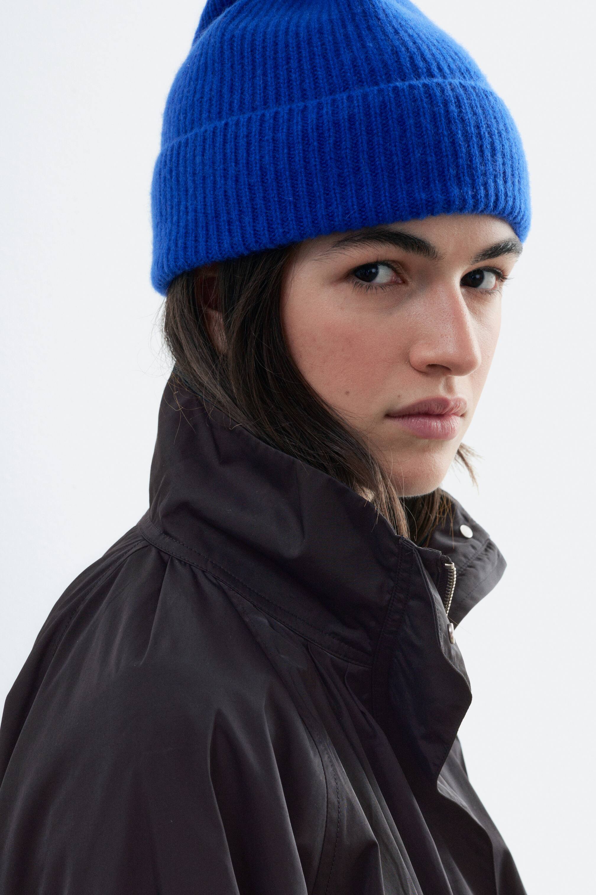 View larger image: A bright blue ribbed knit beanie with a fold-over cuff is being worn by the model, who is also wearing a high-collared black jacket.