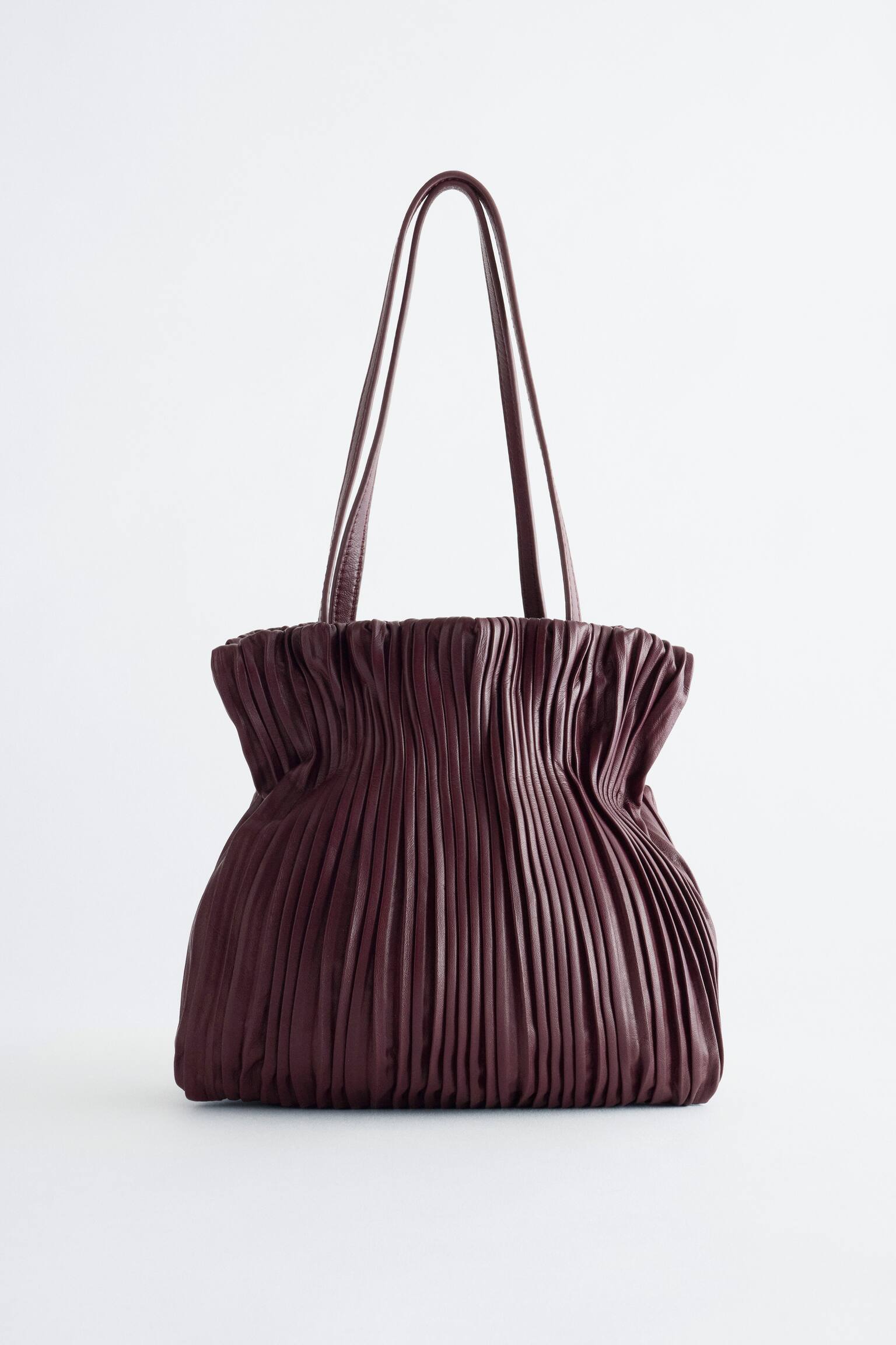Una mini borsa in pelle color bordeaux intenso Ã¨ in posizione verticale in una vista frontale, con una fitta plissettatura verticale su tutto il corpo, che crea una forma stretta nella parte superiore. Due sottili spallacci in pelle abbinati si estendono verso lâalto dallâapertura arricciata.