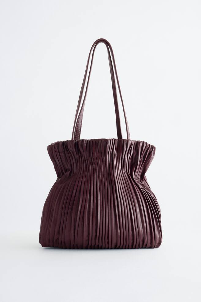 Un bolso mini de cuero burdeos oscuro aparece en posiciÃ³n vertical en una vista frontal, con un fruncido vertical denso que recorre todo el cuerpo y crea una silueta ajustada cerca de la parte superior. Dos finas bandoleras de cuero a juego se elevan desde la abertura fruncida. - 1