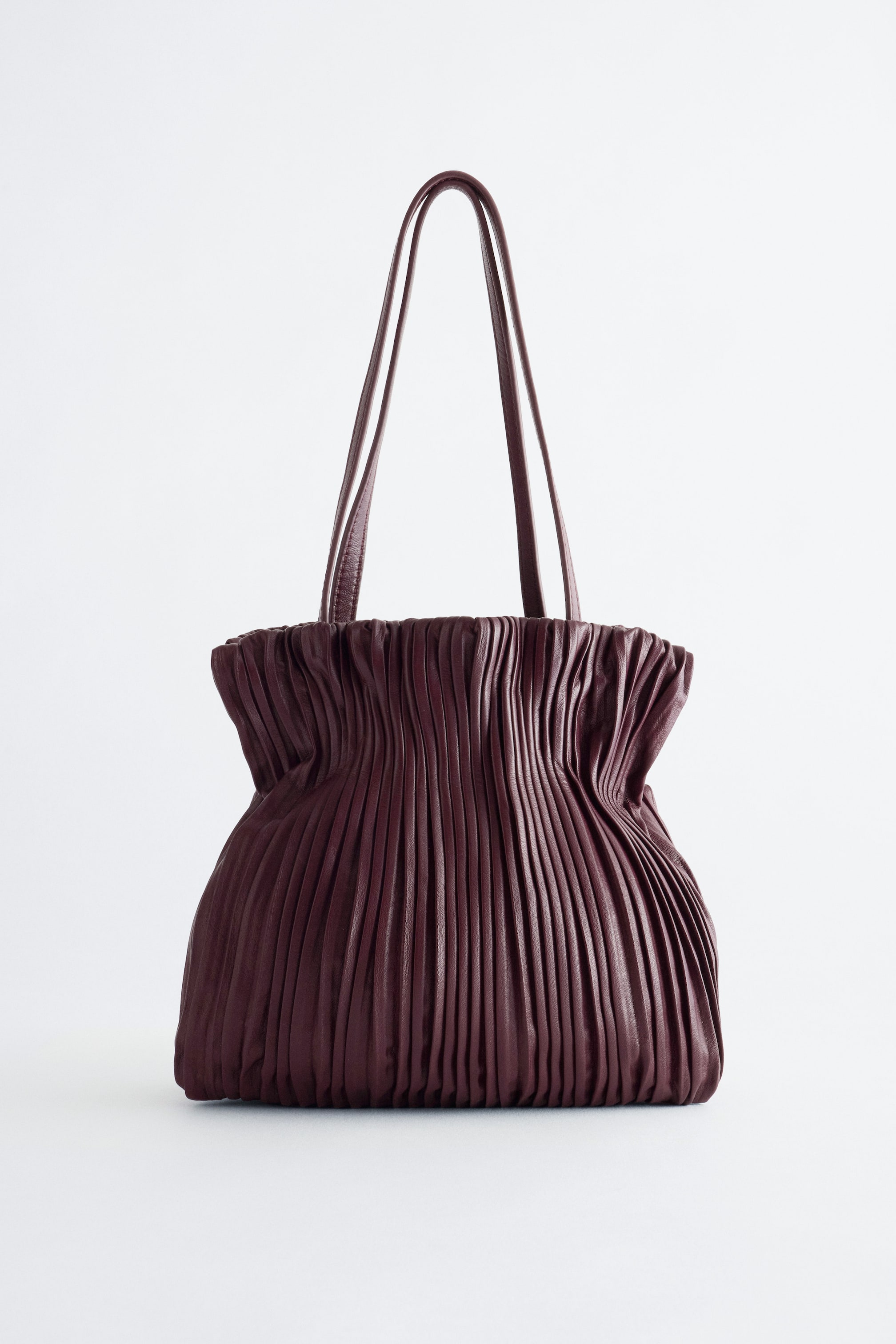 View larger image: A deep burgundy leather mini bag is standing upright in a front view, featuring dense vertical pleating across the body, creating a cinched shape near the top. Two thin, matching leather shoulder straps extend upwards from the gathered opening.