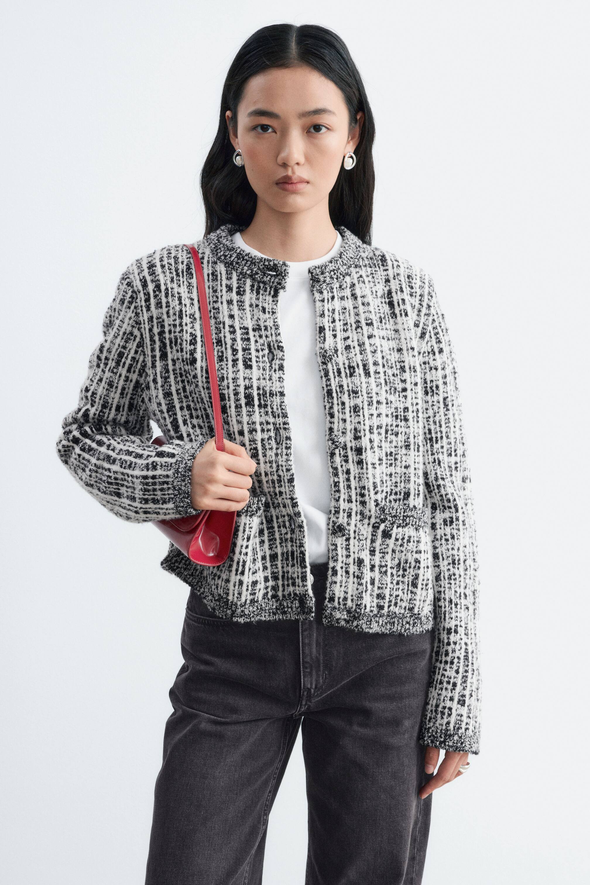 View larger image: A black and white textured knit cardigan featuring structured shoulders and ribbed trims is worn open by the model over a white T-shirt. The outfit is styled with dark grey jeans, and a red shoulder bag is carried under the model’s arm.