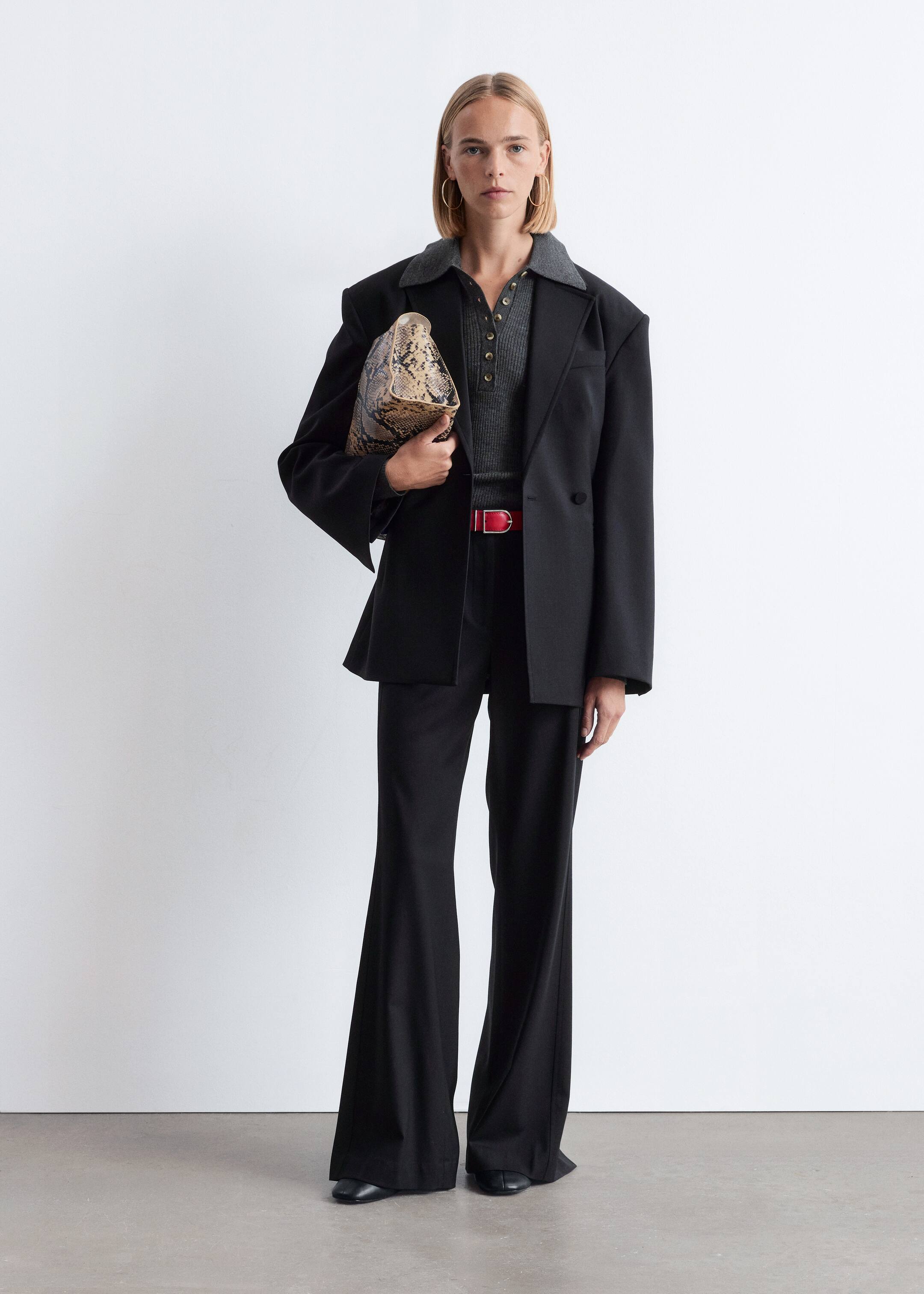 Vis større bilde: Produktet er et ensemble med en svart oversized blazer med hakkeslag, lagd over en grå ribbestrikket poloskjorte med knappestolpe. Sorte bukser med vide ben fullfører antrekket, strammet inn av et rødt belte med et sølvspenne, med en clutchveske i slangeskinnsmønster som holdes og svarte flate sko som er synlige.