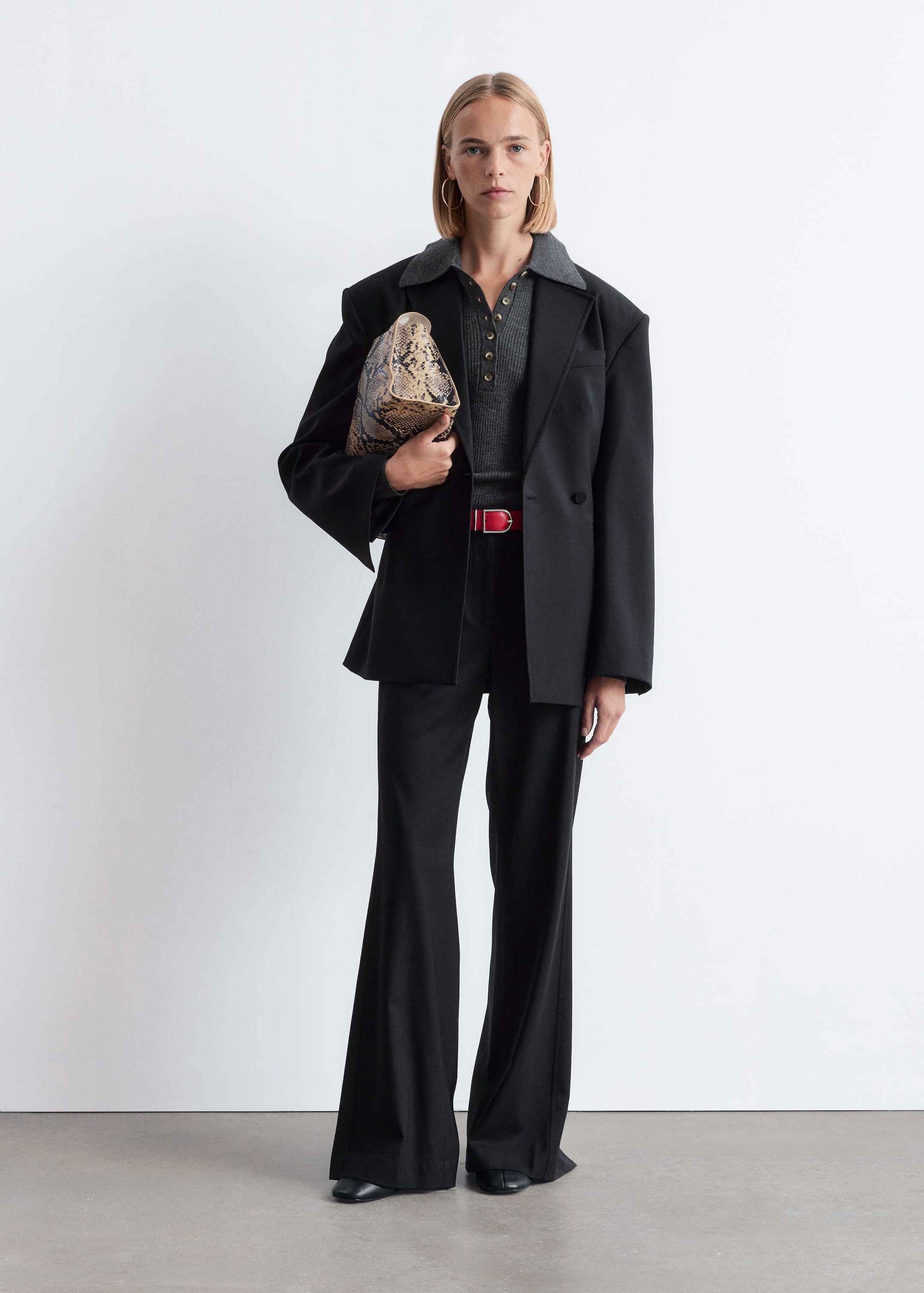 Agrandir l'image: Le produit est un ensemble composé d'un blazer noir oversize avec des revers crantés, superposé à un polo en maille côtelée grise avec une patte de boutonnage. Un pantalon noir à jambes larges complète le look, ceinturé par une ceinture rouge avec une boucle argentée, avec une pochette à motif peau de serpent tenue à la main et des chaussures plates noires visibles.