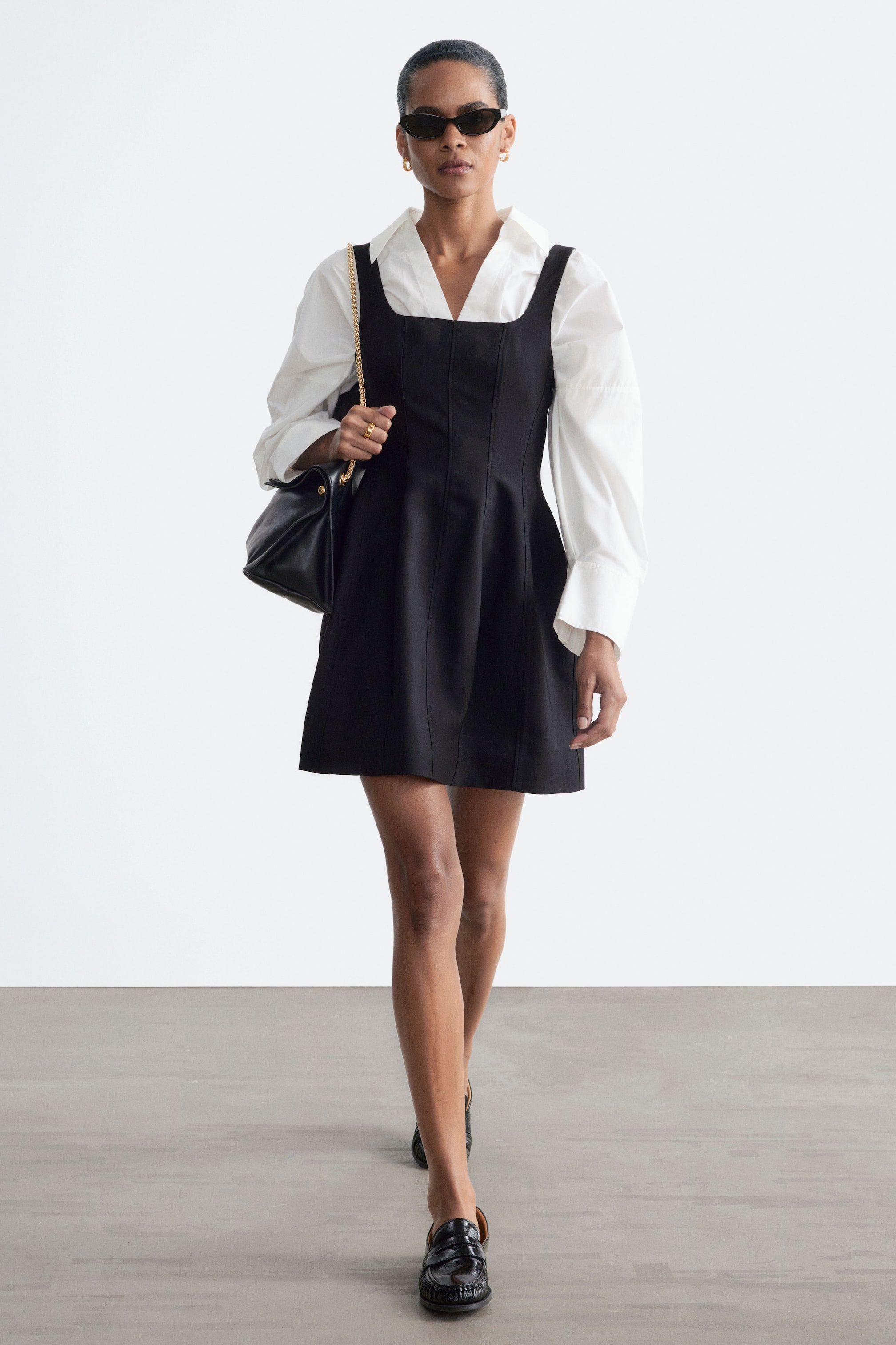 Agrandir l'image: Une mini-robe noire à encolure carrée, avec des coutures verticales surpiquées et une jupe évasée, est portée par le mannequin. La robe est superposée sur une chemise blanche à manches longues et col, et accompagnée d’un sac à bandoulière noir.