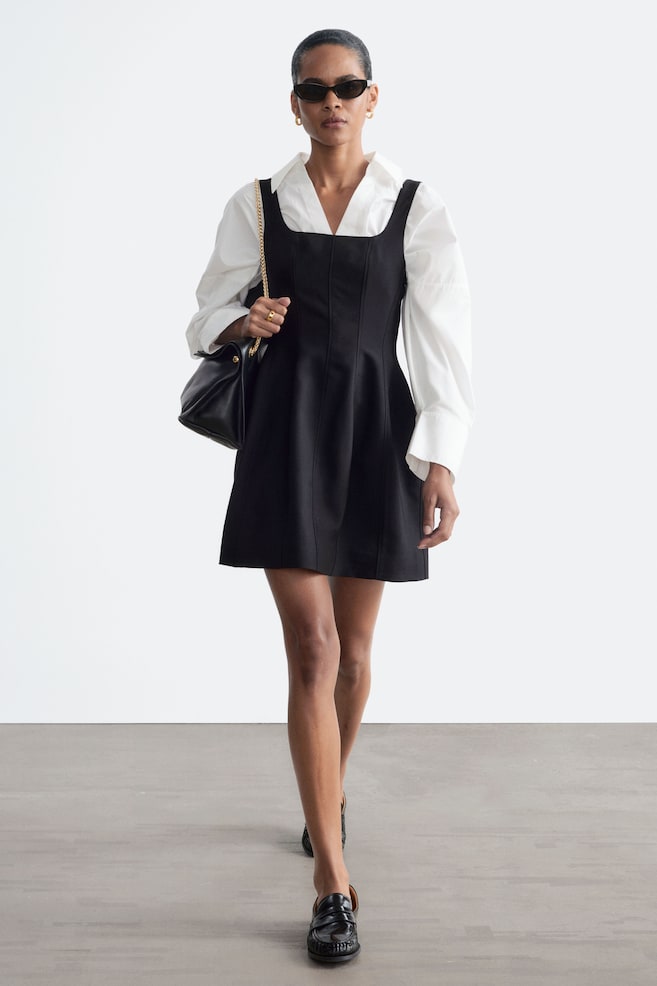 Une mini-robe noire à encolure carrée, avec des coutures verticales surpiquées et une jupe évasée, est portée par le mannequin. La robe est superposée sur une chemise blanche à manches longues et col, et accompagnée dâun sac à bandoulière noir. - 1