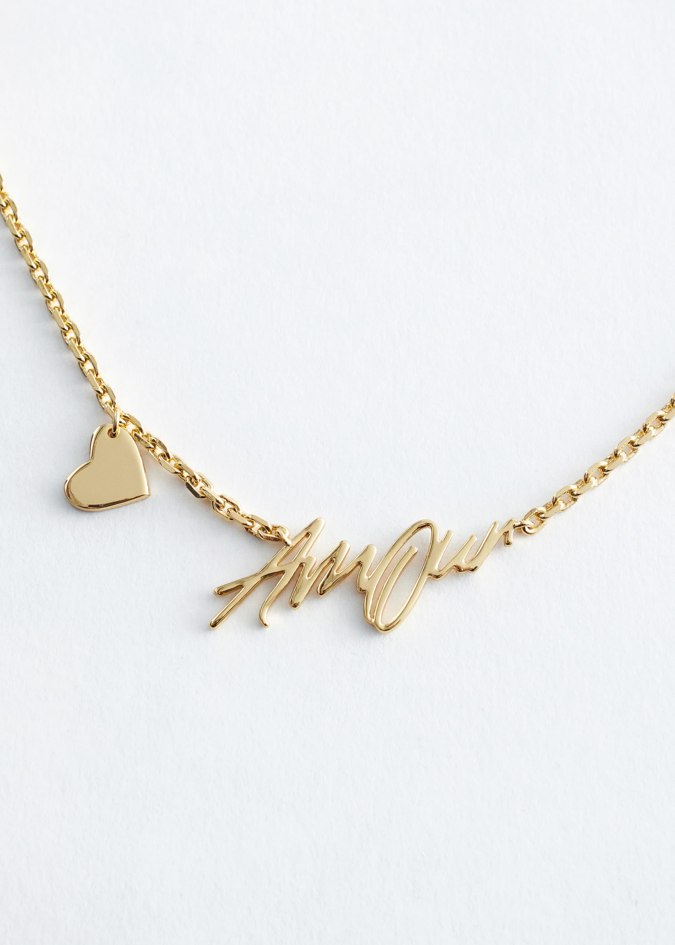 View larger image: A polished brass necklace is featuring a delicate rolo chain, with a statement text-motif pendant spelling "Amour" at the front and a heart charm accenting one side of the chain.