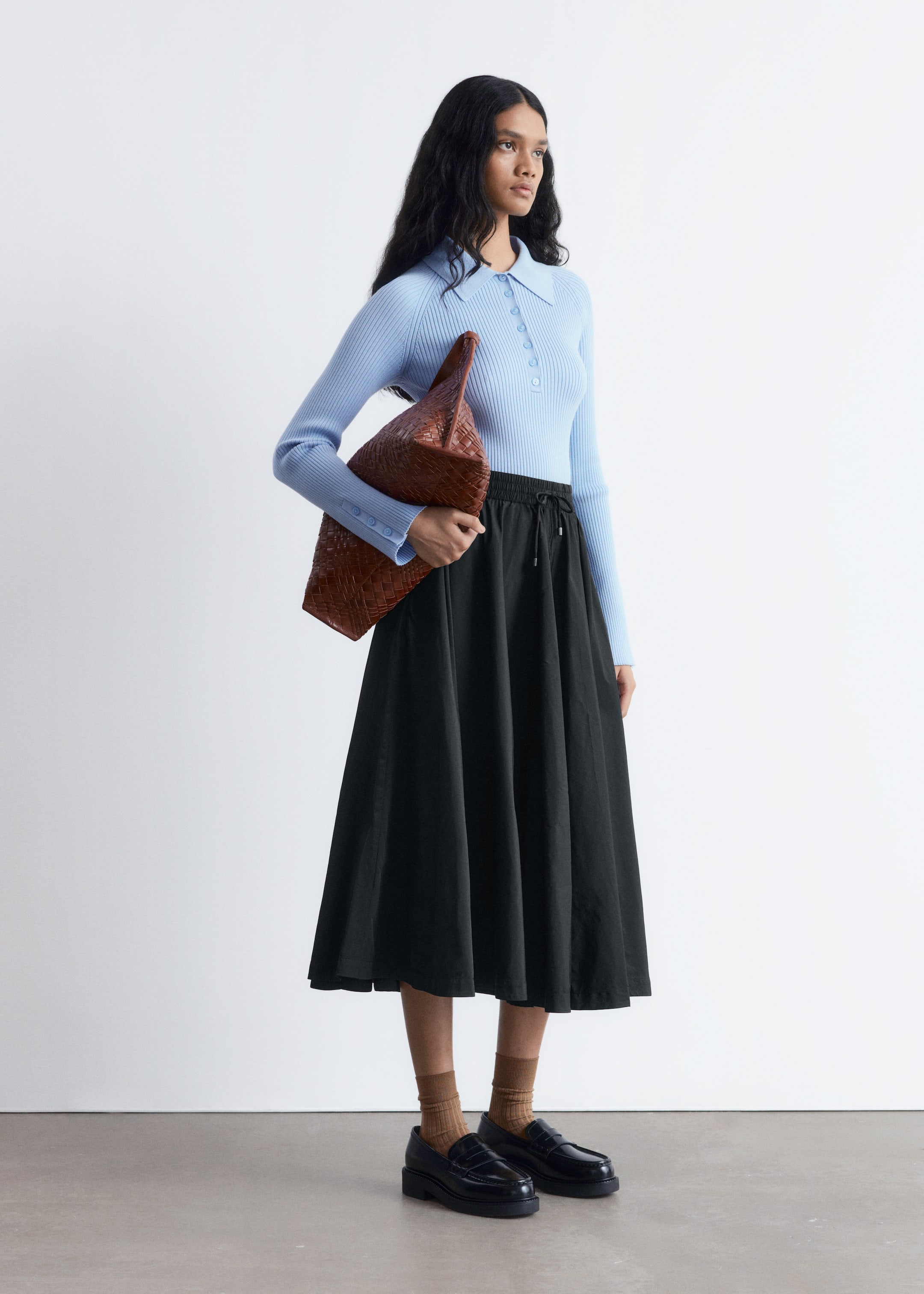 Agrandir l'image: Un haut polo à manches longues bleu clair côtelé, avec un col et une patte de boutonnage, est porté avec une jupe midi noire en ligne A qui a une taille à cordon. Un sac fourre-tout tissé marron est tenu, tandis que des mocassins noirs épais sont portés avec des chaussettes côtelées marron, complétant la tenue.