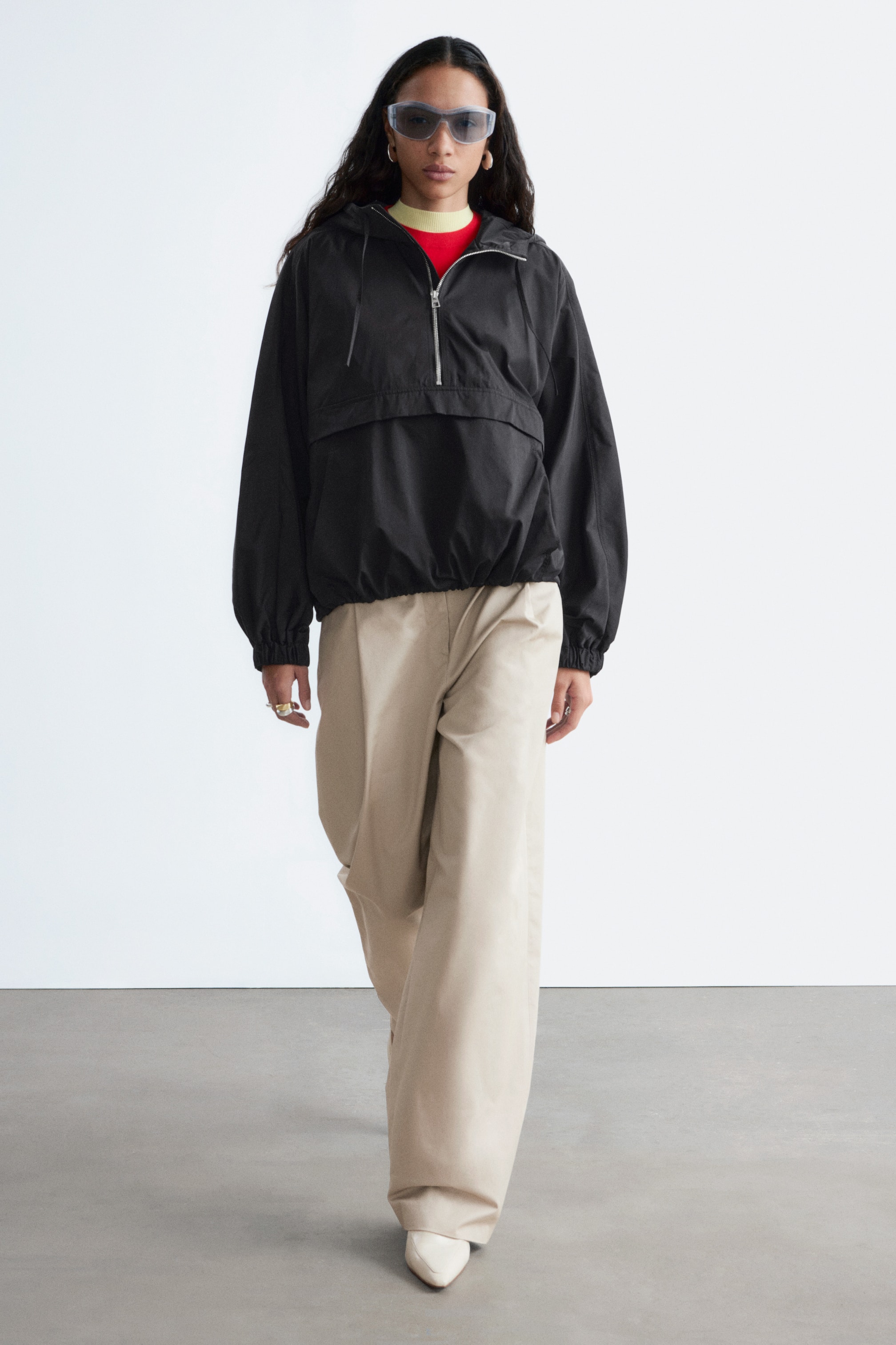 Agrandir l'image: Un anorak noir avec une fermeture éclair argentée à mi-longueur, une capuche avec cordons de serrage visibles, et des poignets et un ourlet élastiqués, superposé à un haut rouge et jaune. Un pantalon beige à jambes larges est également porté, avec un devant plissé et une longueur complète qui tombe sur des chaussures à bout pointu.