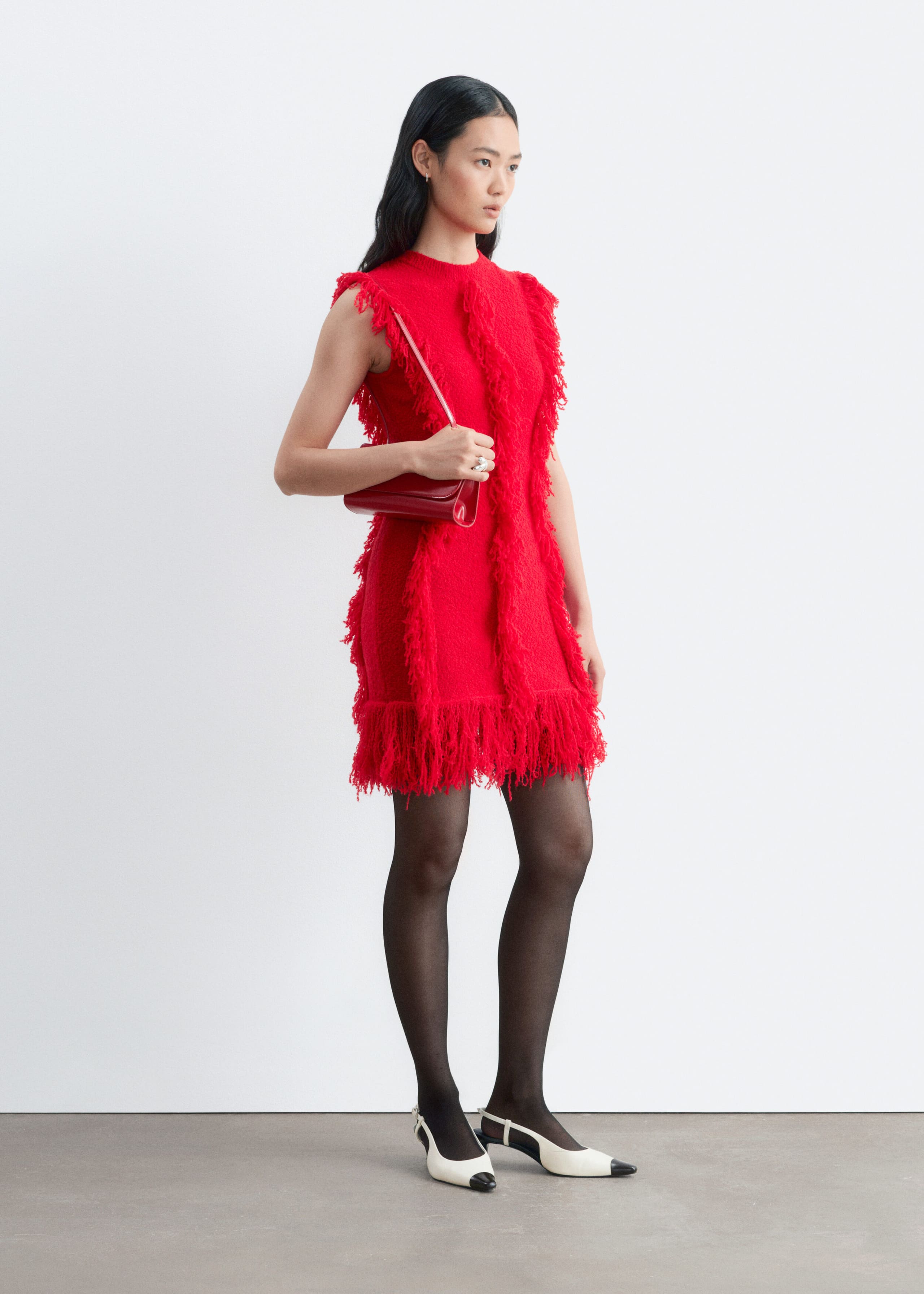 A red sleeveless dress featuring vertical rows of textured fringe and a heavily fringed hem is worn by the model. The model is styling the dress with a matching red shoulder bag, black sheer tights, and white slingback heels with black toe caps.