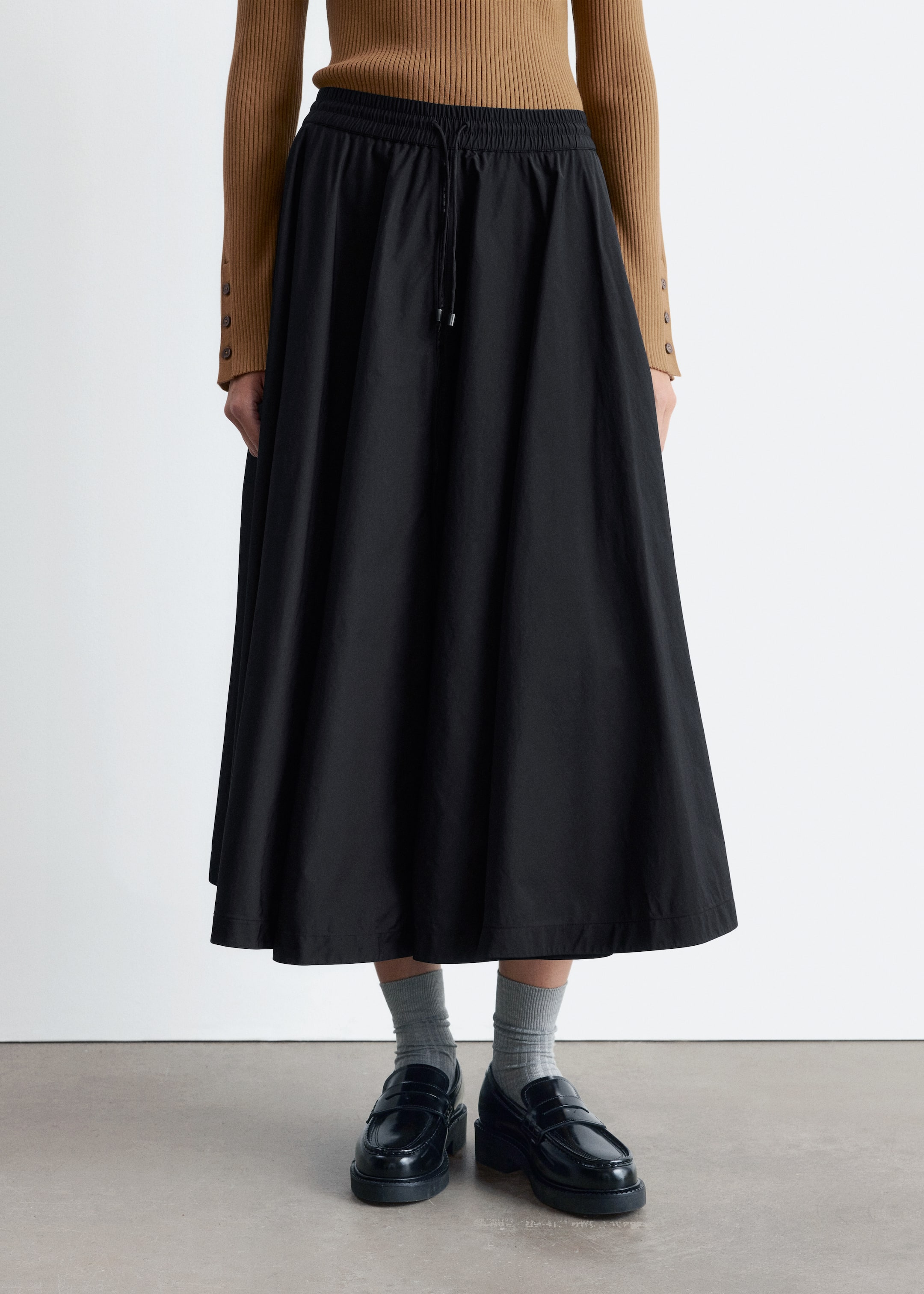 Agrandir l'image: Une jupe midi noire avec une silhouette ample et évasée et une taille élastiquée avec cordon de serrage est portée par le mannequin. Elle est associée à un haut côtelé marron et des mocassins noirs épais.