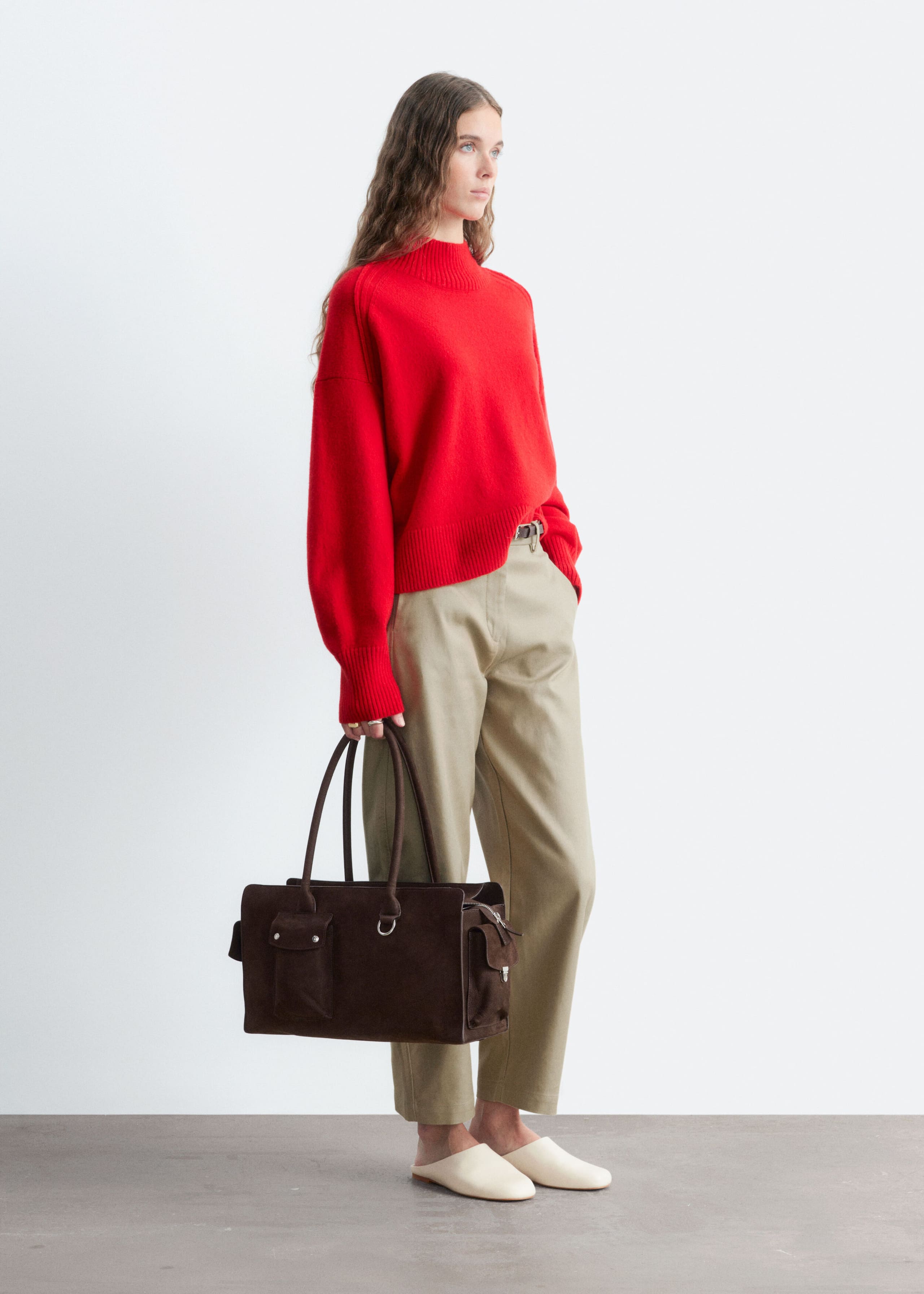 A red mock neck sweater featuring a ribbed collar, cuffs, and hem is being worn by the model. The relaxed-fit knitwear is being paired with beige pants and a dark brown handbag.