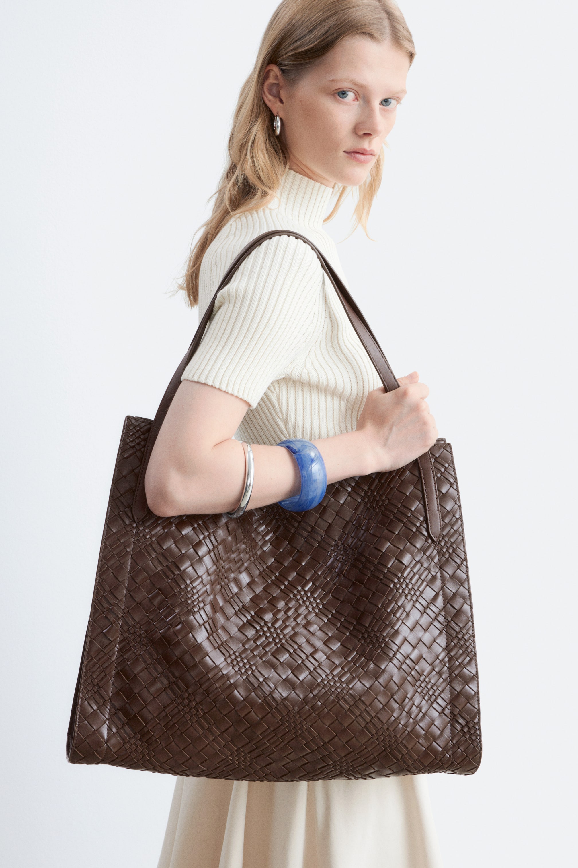 Agrandir l'image: Un sac cabas marron présente un motif tissé entrelacé complexe sur sa face avant, légèrement incliné vers la droite. Il est tenu par une personne, avec une bandoulière plate reposant sur son épaule.