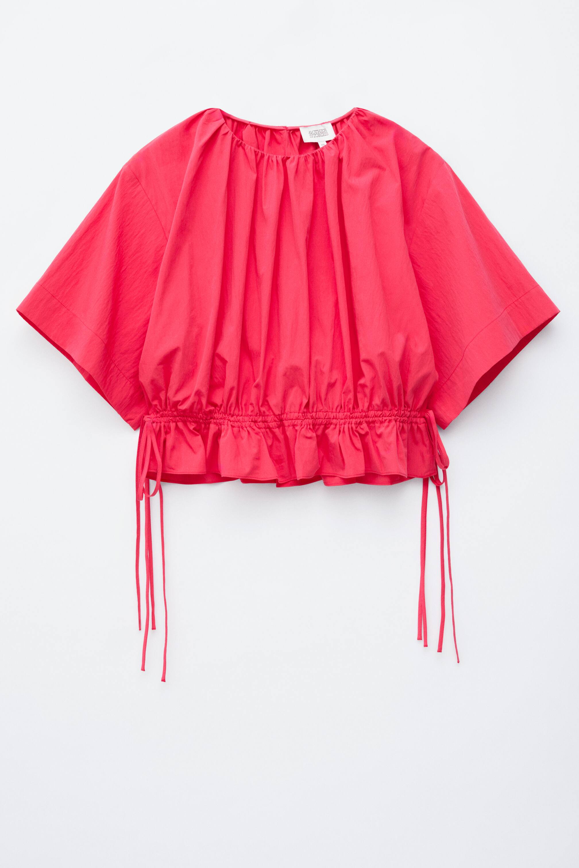 Zobrazit větší obrázek: Živý červený top s krátkým rukávem je zobrazen zepředu, s řasením u kulatého výstřihu. Tunýlek se šňůrkami v pase vytváří přiléhavý siluetu, s malým volánkem podél spodního okraje.