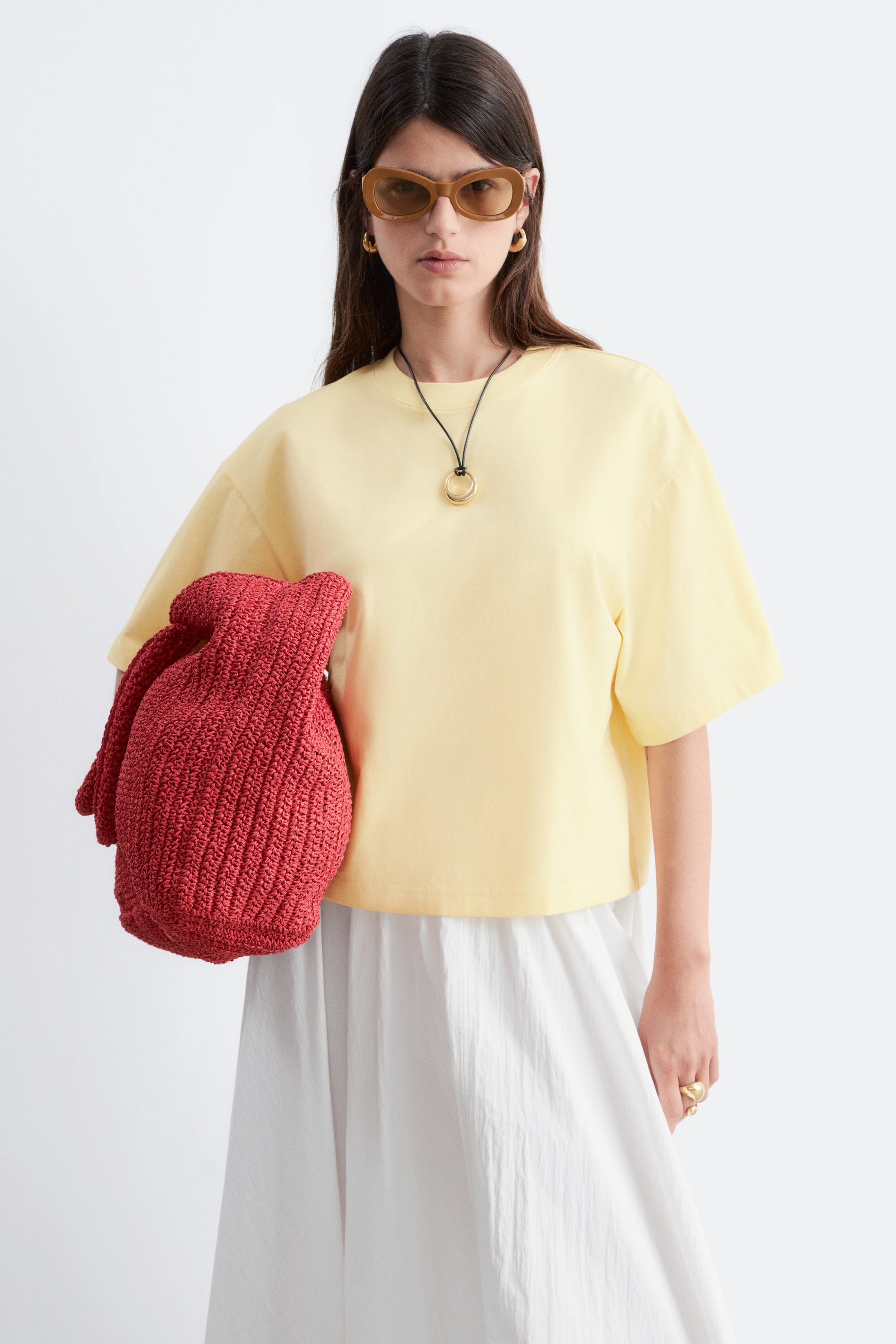 Zobrazit větší obrázek: Světle žluté tričko s kulatým výstřihem a krátkými, volnými rukávy. Nabízí uvolněný, mírně zkrácený střih, vrstvený přes bílou sukni. Tmavý šňůrkový náhrdelník zdobí jeho výstřih.