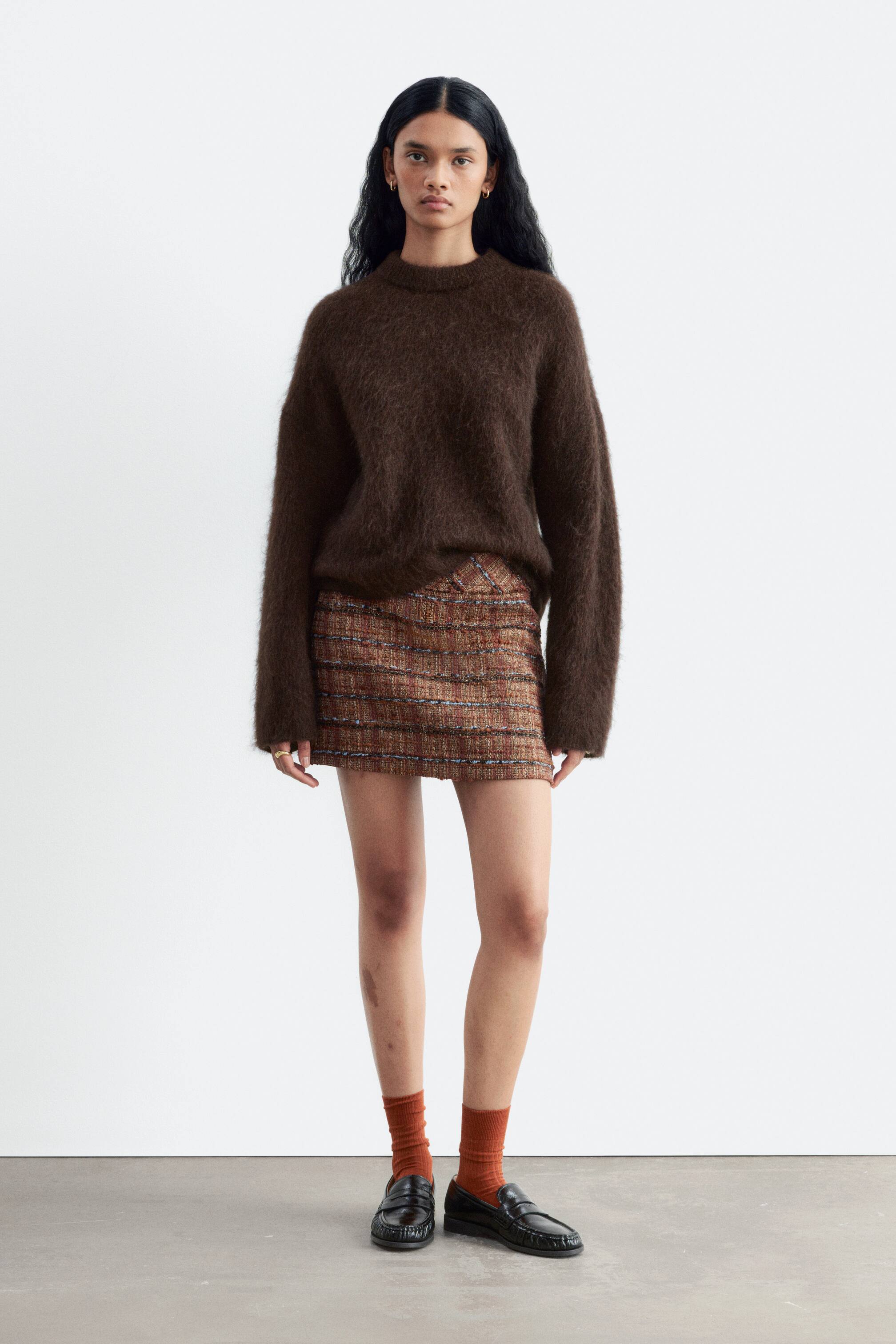 Visualizza l'immagine ingrandita: Una minigonna in tweed con un motivo testurizzato marrone a righe orizzontali indossata dalla modella. La gonna è abbinata a un morbido maglione a maniche lunghe color marrone scuro drappeggiato sulla cintura, calzini color ruggine e mocassini neri.